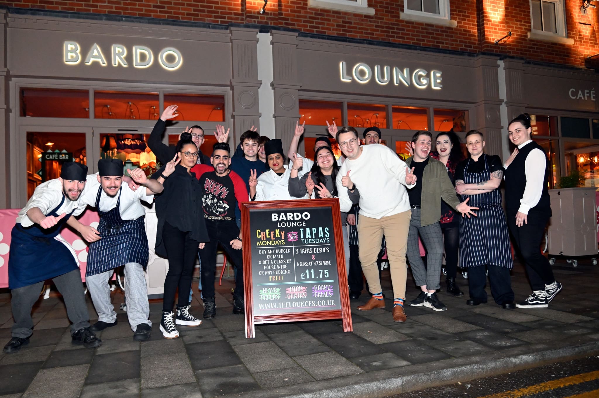 A group of cheerful staff pose together outside Bardo Lounge restaurant at night, surrounding a signboard advertising Bardo deals. The warmly lit building and café sign glow in the background.