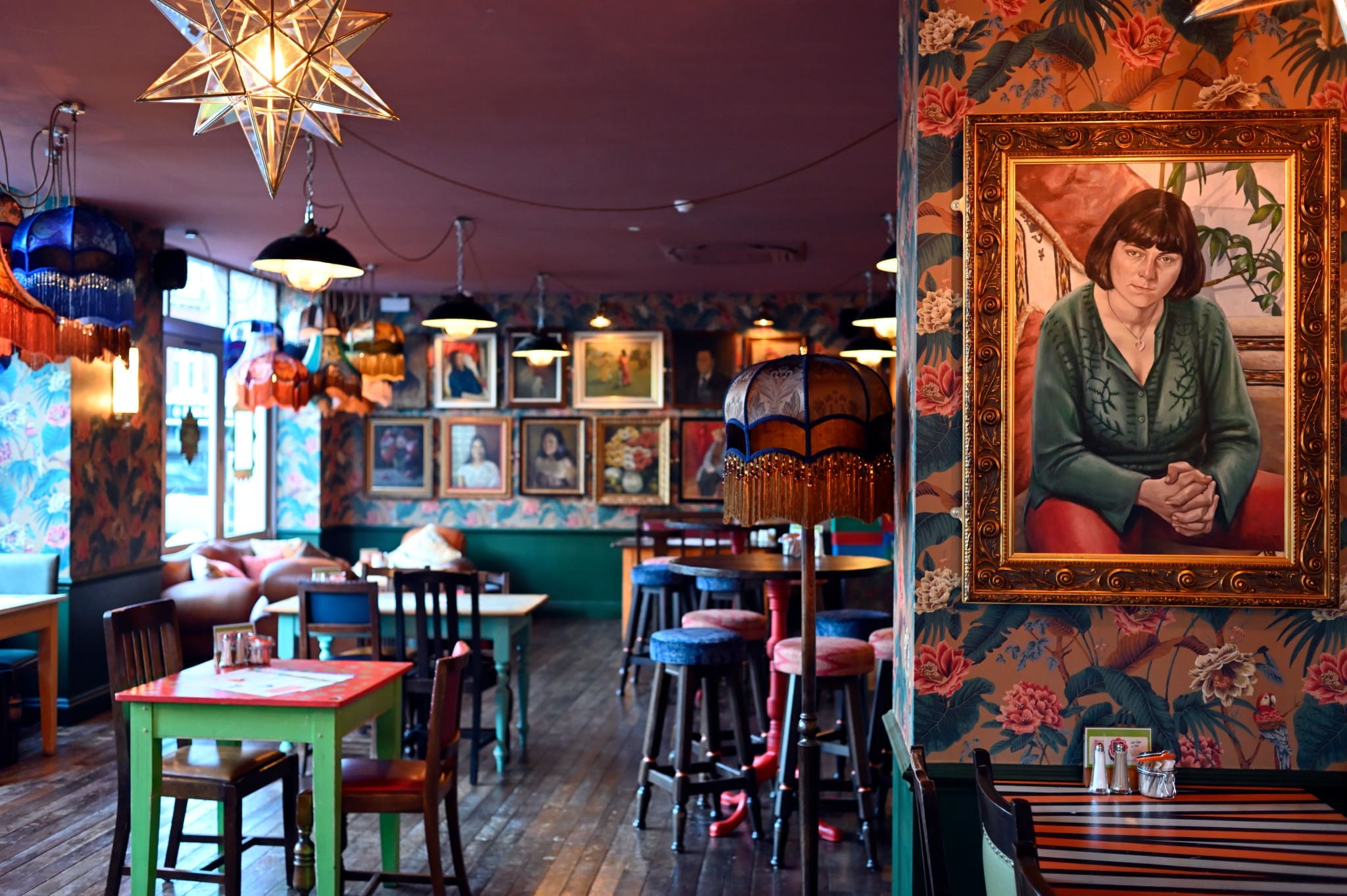 A cozy, vintage-style café with floral wallpaper, eclectic lampshades, wooden tables, and numerous framed portraits on the walls evokes a Bardo-like atmosphere. A large portrait of a woman in green is prominently displayed in the foreground.