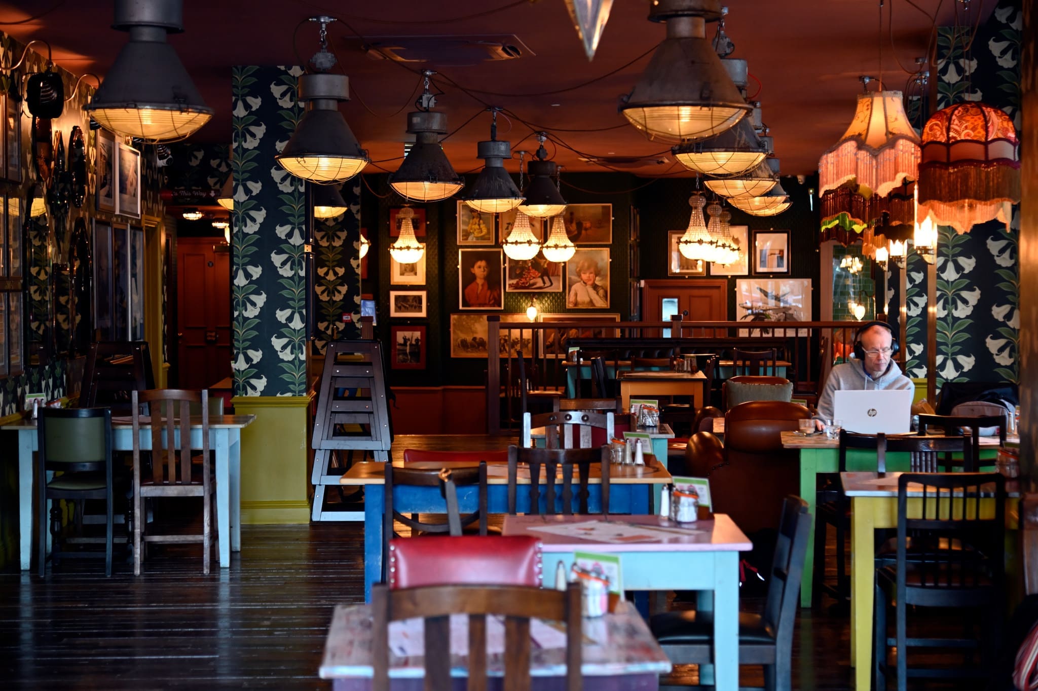 A cozy, dimly lit café called Bardo with colorful mismatched chairs and tables, vintage lamps, framed photos on the walls, and one person sitting alone using a laptop. The space feels inviting and quiet.