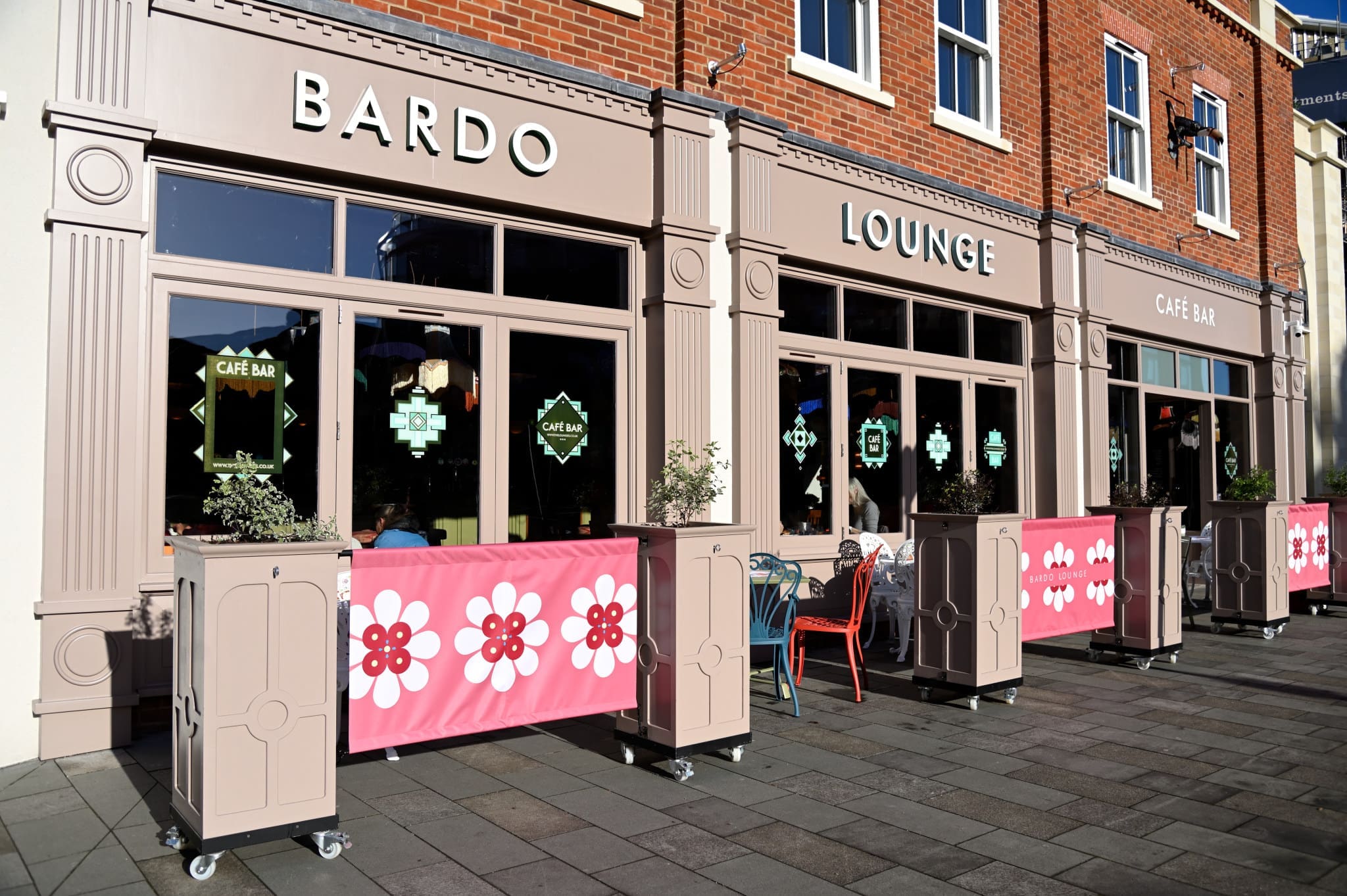 The exterior of Bardo Lounge café bar features large windows, pink barriers adorned with white and red flowers, and outdoor tables and chairs set on a sunlit paved sidewalk, inviting guests to experience the vibrant Bardo atmosphere.