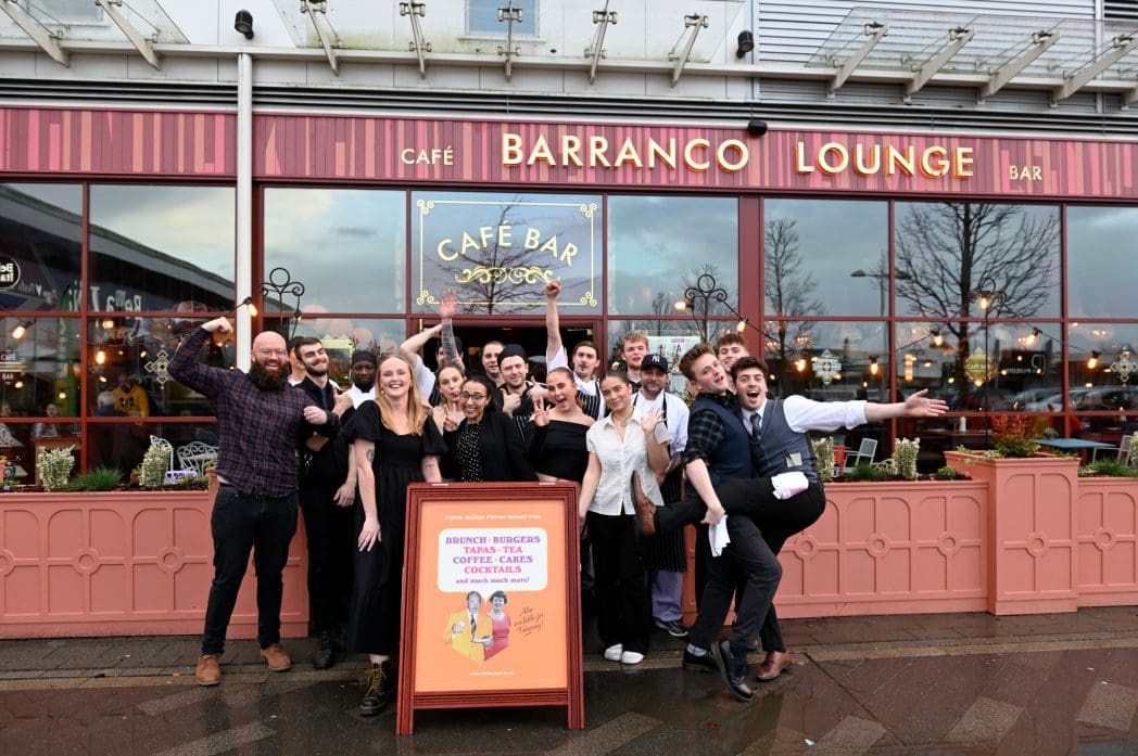 A group of smiling people stand together in front of Barranco Lounge Café Bar, posing cheerfully with arms raised around a large Barranco sign promoting “Brunch, Burgers, Sharing, Coffee, Teas, Cocktails.”.
