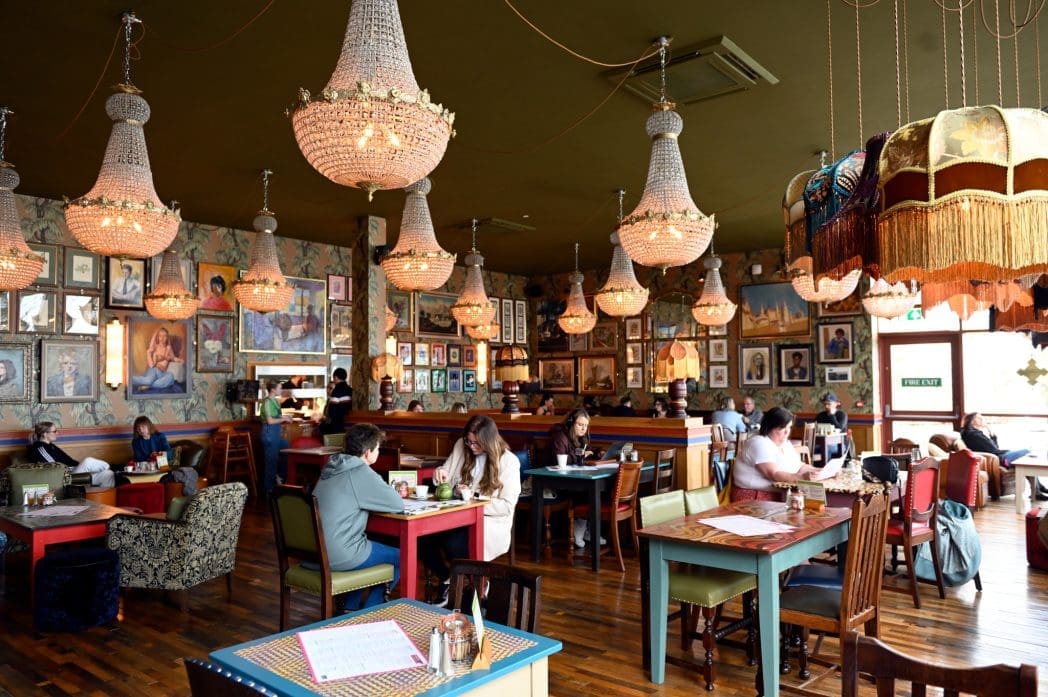 People dine and chat at colorful tables in a cozy Barranco restaurant decorated with vintage chandeliers, eclectic lamps, and framed pictures on patterned wallpaper. The wooden floor adds warmth to the lively atmosphere.