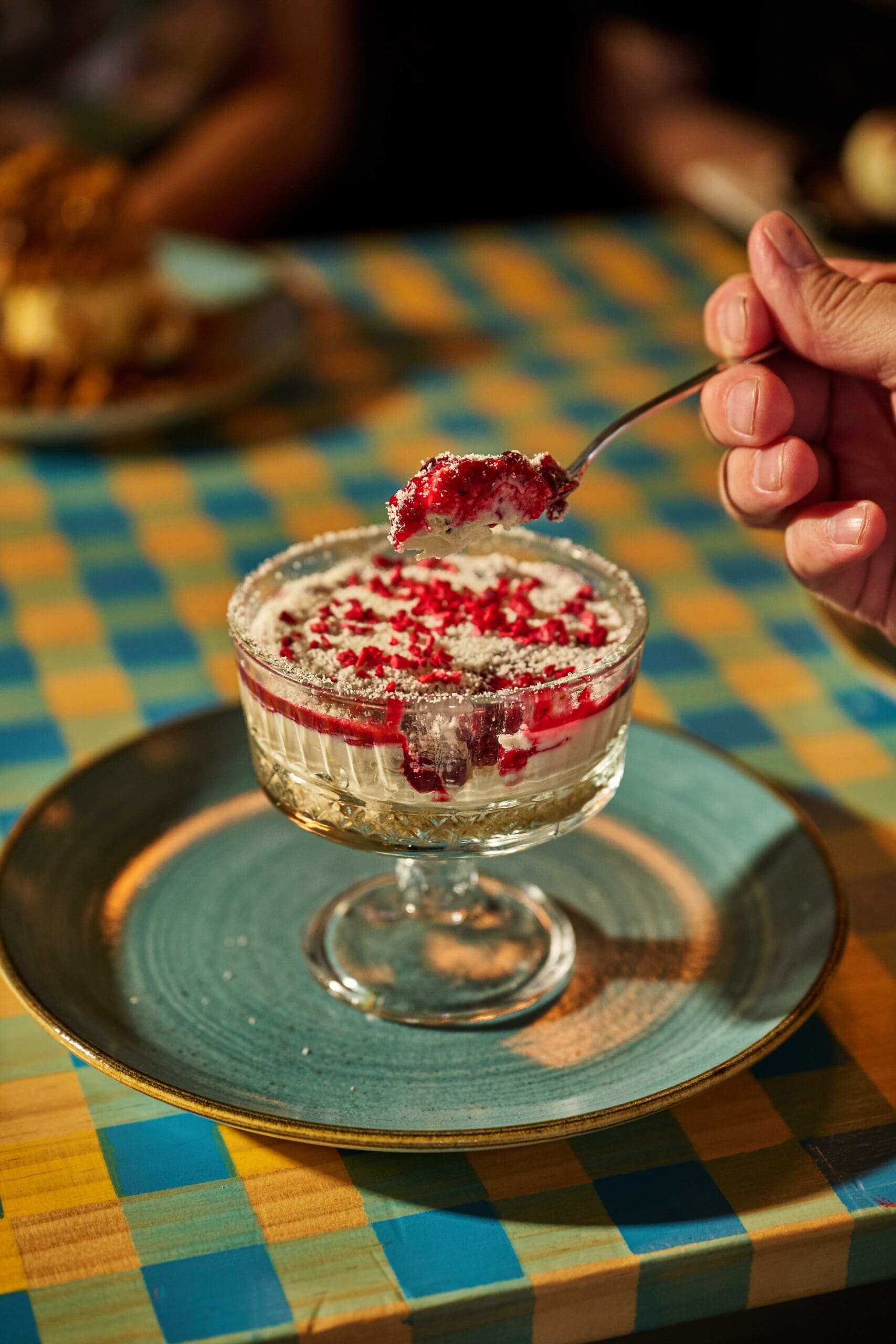 A hand holds a spoon with a bite of dessert above a glass dish of creamy dessert topped with red berry pieces, set on a blue plate atop a checkered tablecloth.