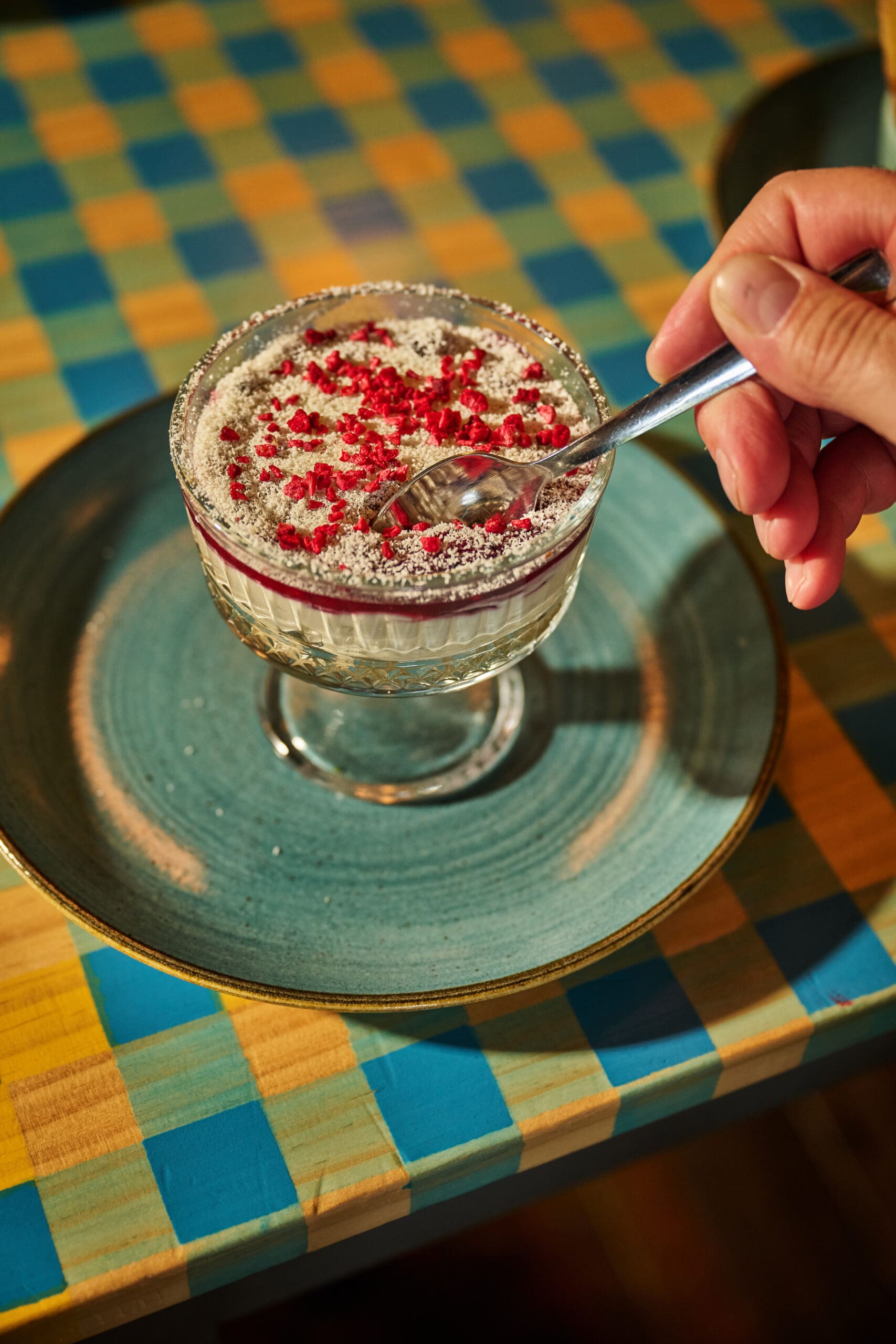 A hand holds a spoon, poised over a glass dessert dish filled with a creamy, layered dessert topped with powdered sugar and red berry bits, set on a blue-green plate on a checkered table.