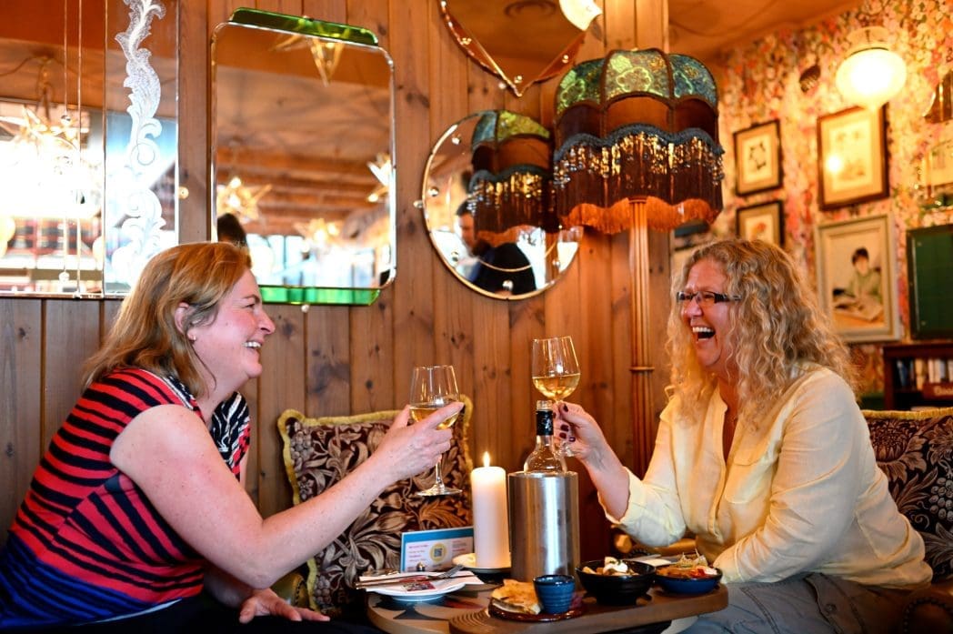 Two women sit in a cozy, vintage-style room, smiling and toasting with glasses of Bianco. The table holds snacks, a lit candle, and a wine bottle. Warm lighting and retro decor create a cheerful, relaxed atmosphere.