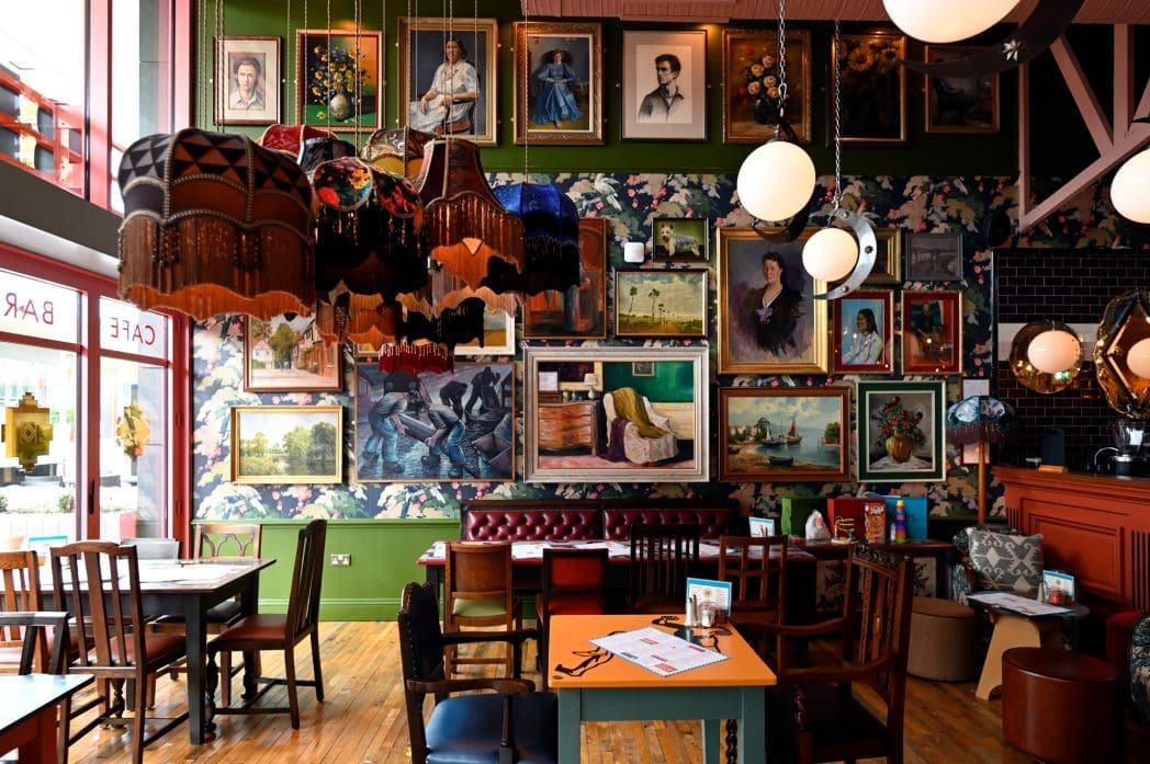 Colorful cafe interior with eclectic vintage decor, mismatched chairs, vibrant Bianco lamps, and a wall covered in framed paintings and portraits. Warm lighting and wooden floors create a cozy, artistic atmosphere.