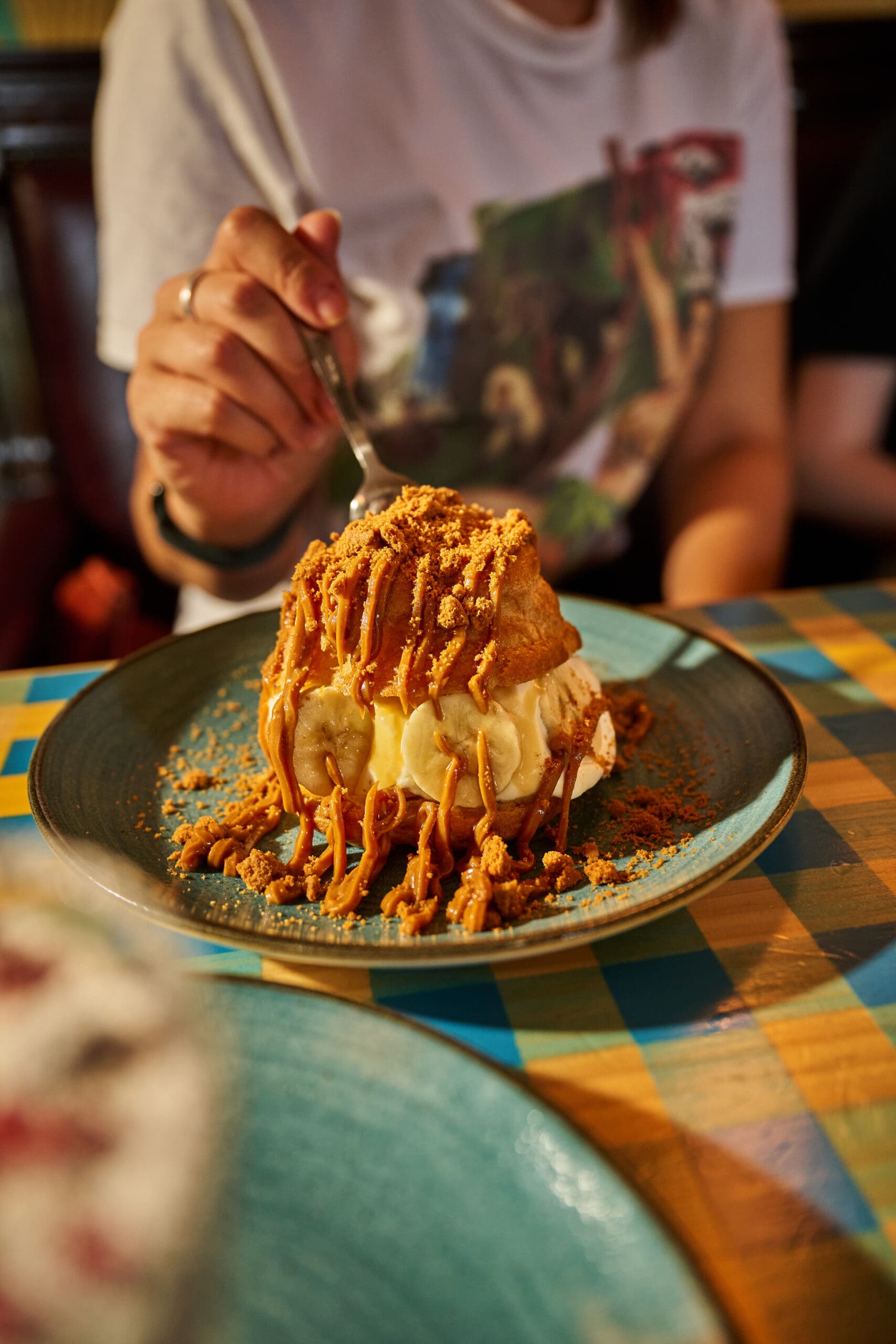 A person holds a fork, about to eat a dessert with ice cream, pastry, caramel sauce, and crushed cookies on a blue plate at a table with a checkered tablecloth.