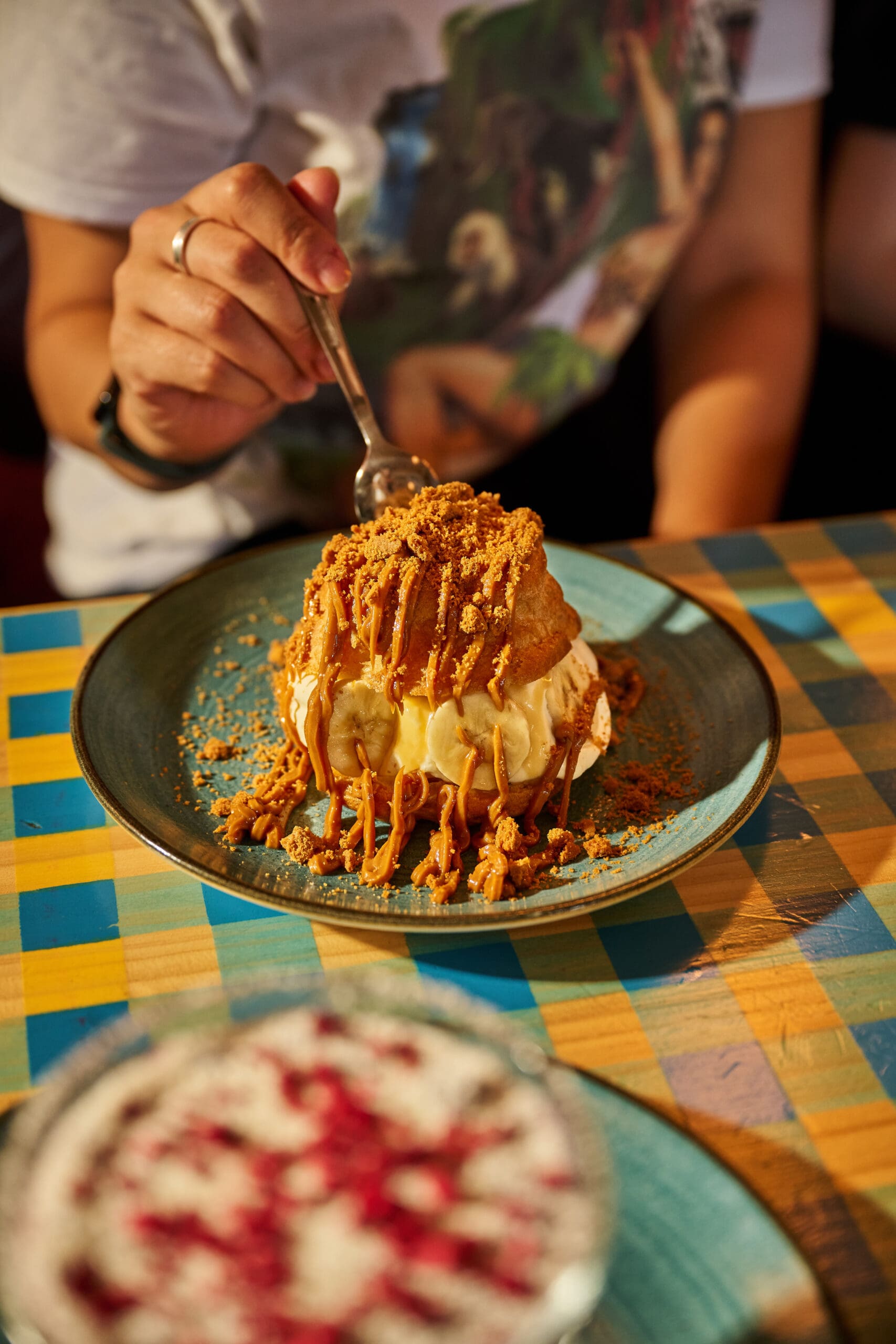 A person holding a spoon is about to eat a dessert topped with crumbled cookies and caramel sauce on a blue plate, set on a colorful checkered table.
