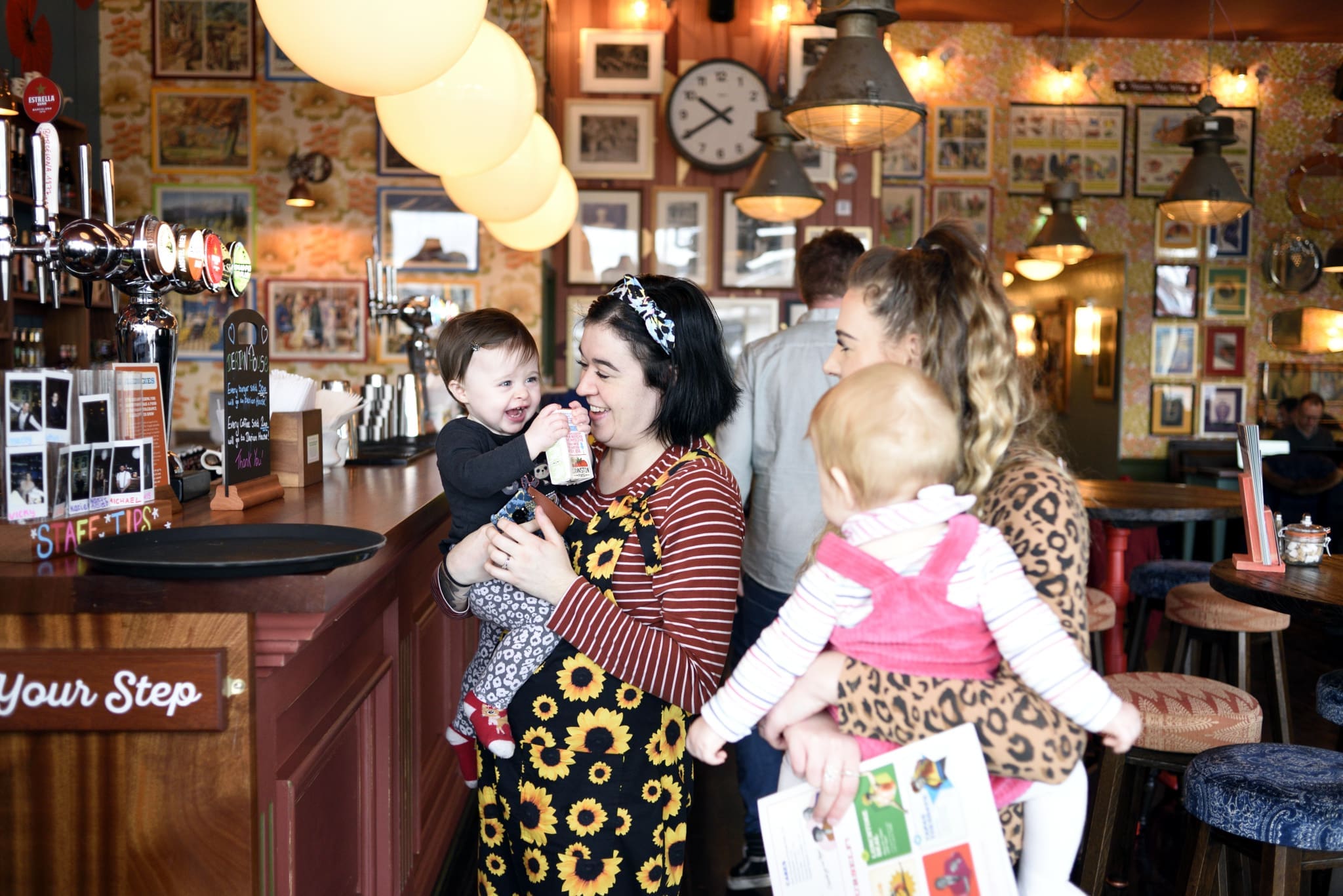 Two women holding babies smile and talk at a cozy bar with calico-patterned wallpaper, framed pictures, and warm lighting. One wears a sunflower dress; the bar features a “Mind Your Step” sign and a clock on the wall.