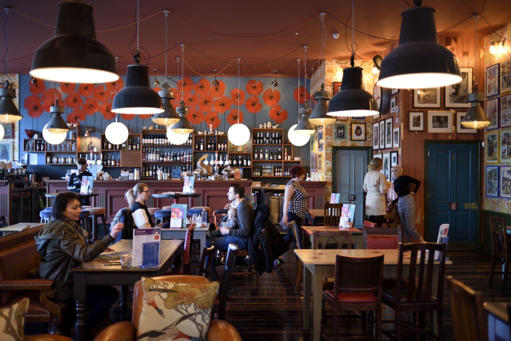A cozy café with people sitting at tables, chatting and drinking. Warm calico lighting hangs from the ceiling, with eclectic wall art and shelves of bottles behind the bar. The atmosphere is lively and inviting.