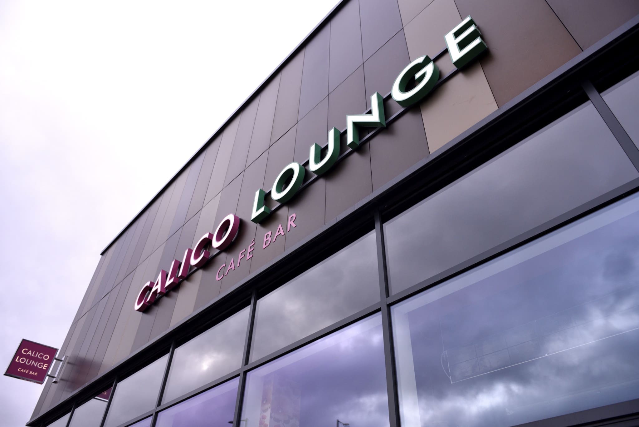 Exterior of a modern building with large windows reflecting the sky. The sign reads “Calico Lounge Café Bar” in bold letters, with “Calico” in pink and “Lounge” in green.
