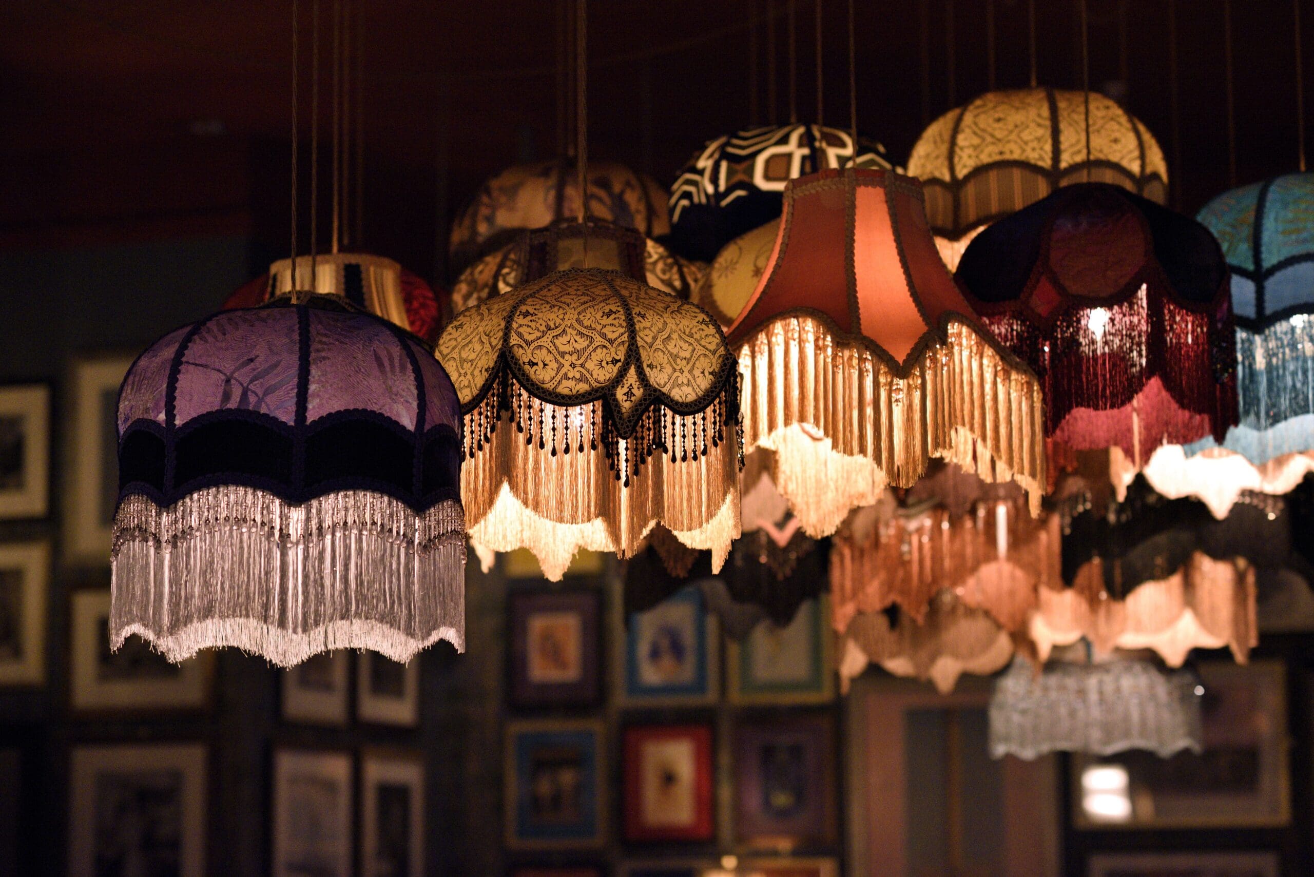 A group of vintage, fringed lampshades from Cassio in various colors and patterns hang from the ceiling, warmly illuminating a room decorated with framed pictures on the walls in the background.