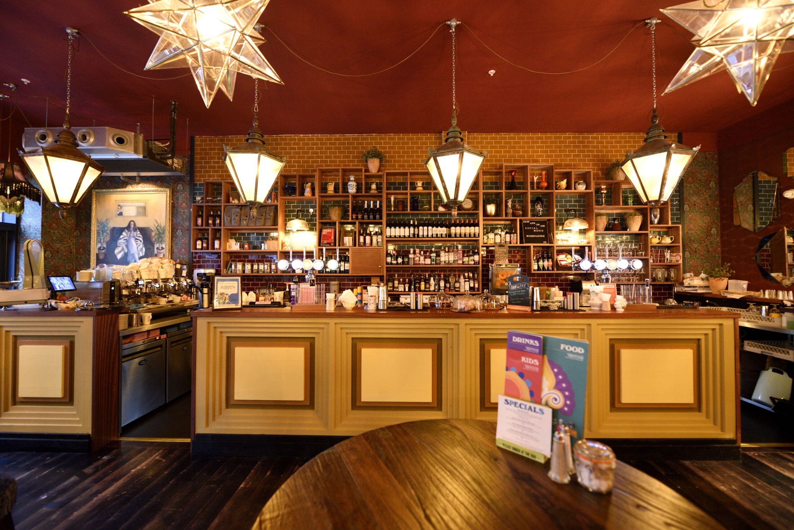 A warmly lit bar with vintage-style hanging lights, Cassio star-shaped ceiling fixtures, a wooden counter, shelves filled with bottles, and food and drink menus displayed on the counter.