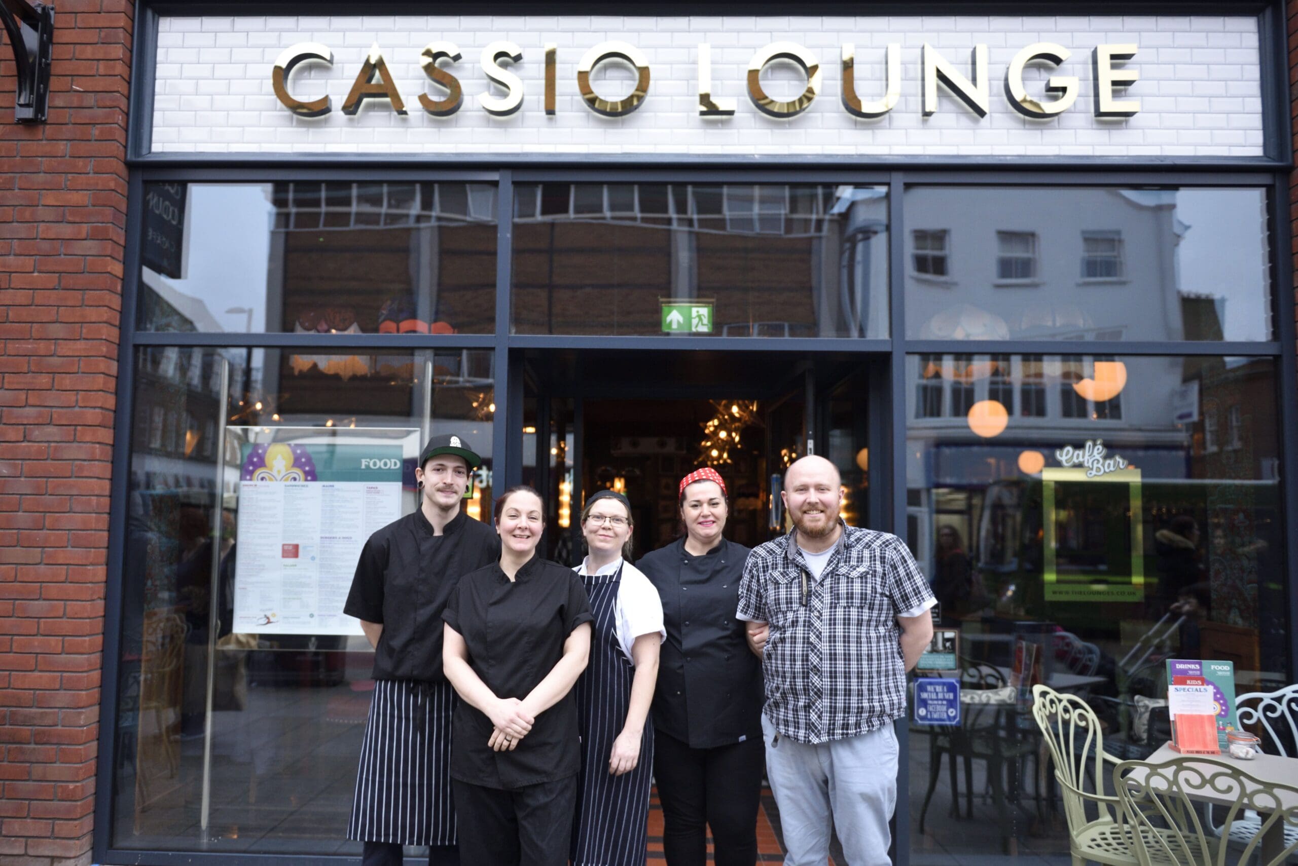 Five people, likely Cassio restaurant staff, stand smiling outside the Cassio Lounge. The group includes men and women in black uniforms and casual clothes, posing cheerfully in front of the restaurant’s glass entrance.