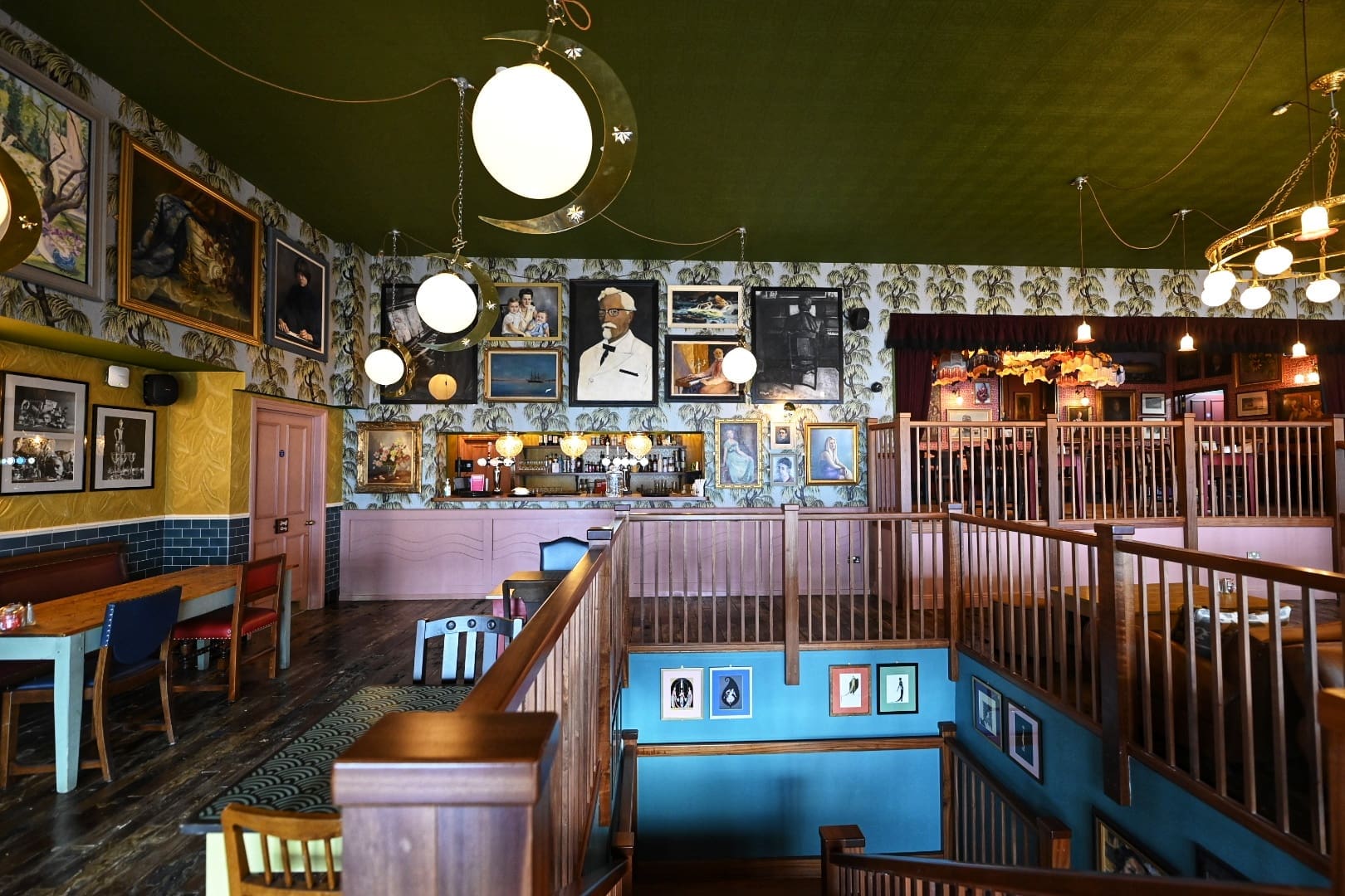 A cozy, eclectic restaurant interior with wooden railings, colorful walls, vintage-style furniture, and a Martello bar area. Numerous framed portraits and photos decorate the walls, including a large image of Colonel Sanders.