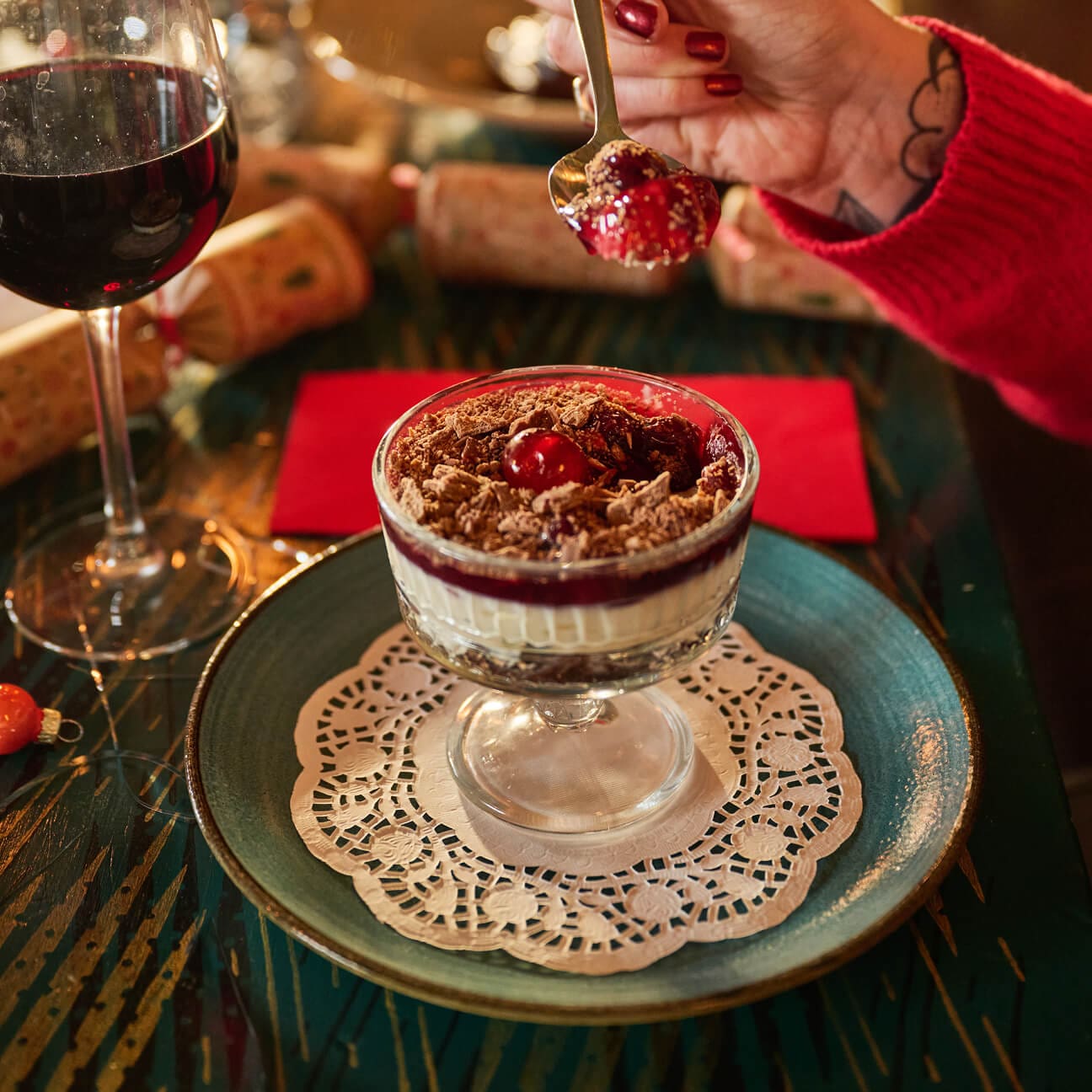 A person in a red sweater holds a spoonful of layered dessert with cherry and chocolate in a glass dish, placed on a blue plate with a lace doily. A glass of red wine and a red napkin are nearby.