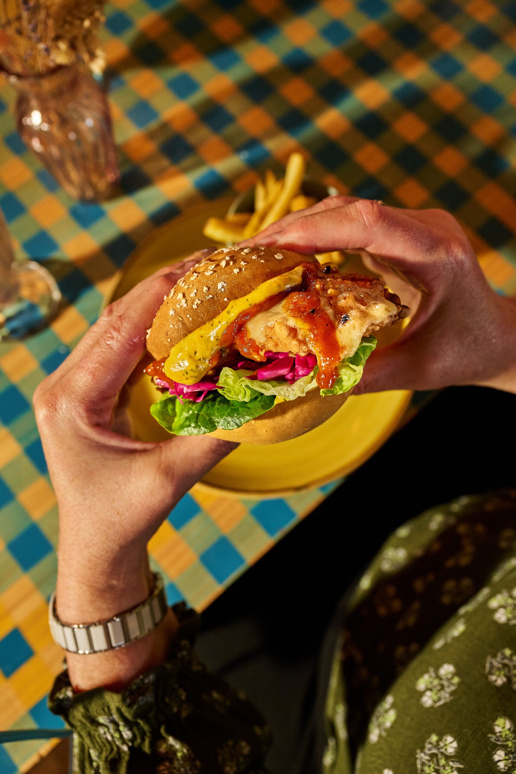 A person holding a chicken sandwich with lettuce, red cabbage, and sauce in both hands over a yellow plate, with fries and a checkered tablecloth in the background.