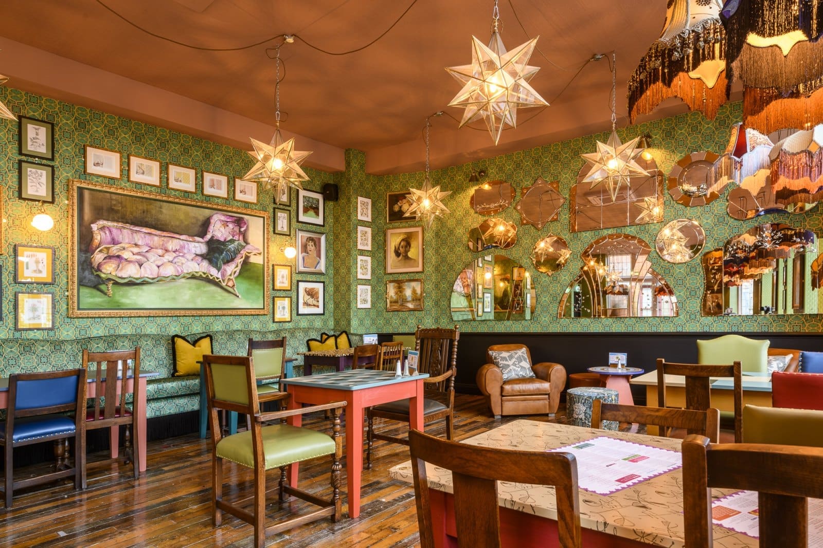 A cozy, eclectic café interior with vintage furniture, patterned wallpaper, Bordo accents, star-shaped pendant lights, framed art, mirrors, and colorful chairs creates a warm, inviting, and artistic atmosphere.