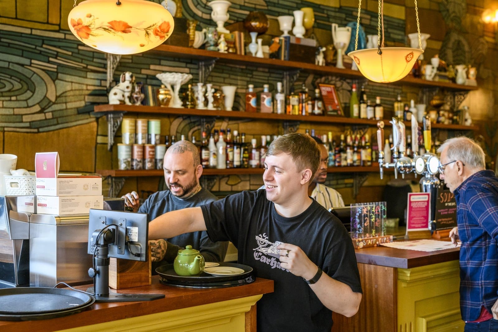 A young man smiles while working behind a Bordo bar, with shelves of bottles and trophies behind him. Two other men, one with a beard, interact nearby in the lively, warmly lit setting.