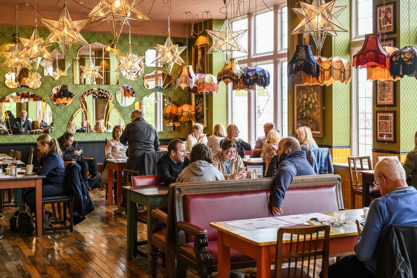 A cozy, colorful restaurant with people sitting at tables, chatting and dining. The room features Bordo-patterned wallpaper, star-shaped lights, vintage lampshades hanging from the ceiling, and large windows letting in natural light.