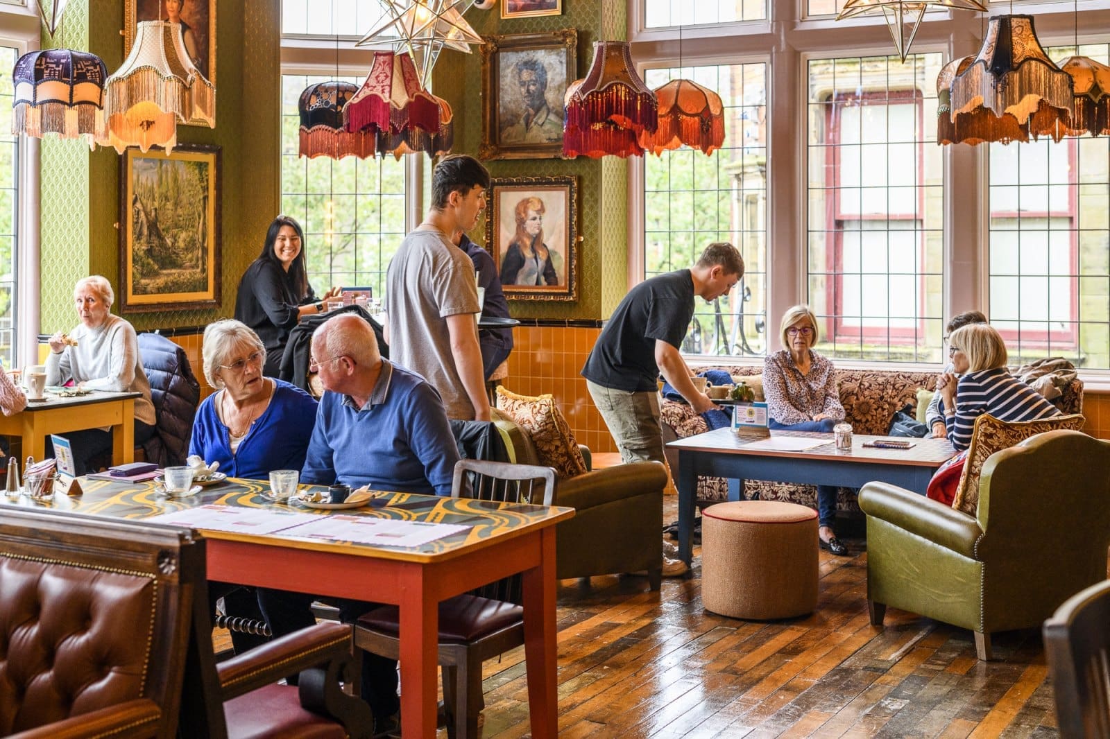 People sit and chat at tables in a cozy, vintage-style cafe with large windows, Bordo-patterned lampshades, and wooden floors. Two staff members serve guests, and framed paintings decorate the eclectic walls.