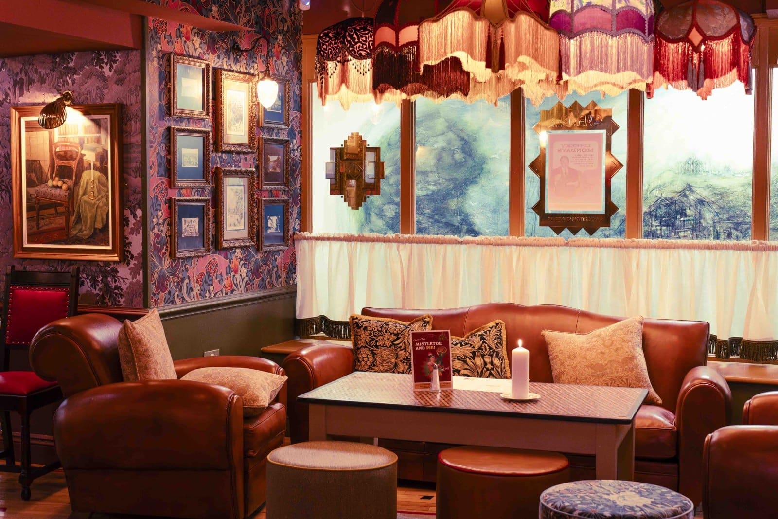 A cozy vintage Bruto lounge with leather armchairs, a sofa, framed pictures on patterned wallpaper, decorative pillows, a candle on the table, and warm lighting from a fringed lamp and wall sconces.