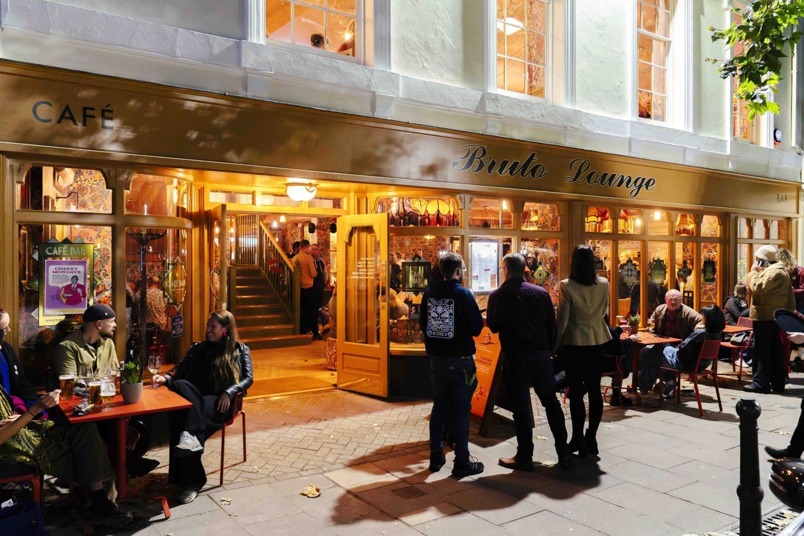 People sitting and standing outside Bruto Lounge at night, enjoying food and drinks at red tables on the sidewalk; large windows reveal the cozy, inviting Bruto interior.