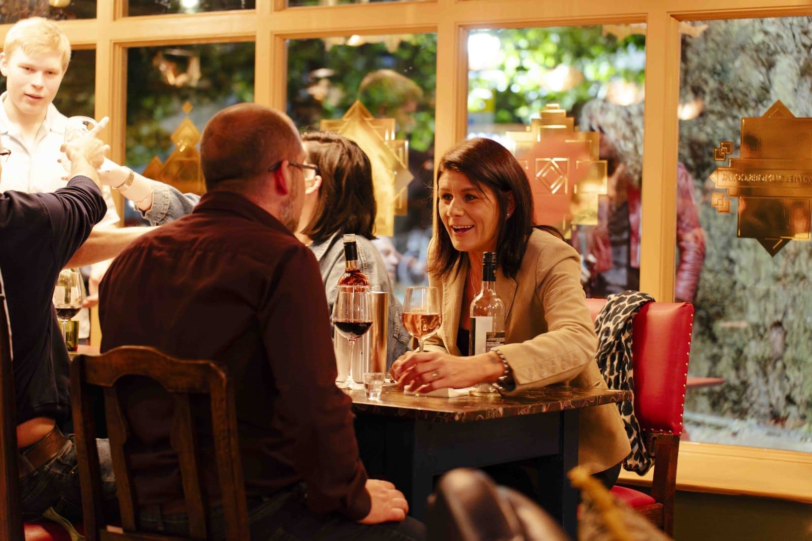 A woman and a man sit at a restaurant table, engaged in conversation over glasses of Bruto wine. Other people are in the background, and warm lighting creates a cozy atmosphere.