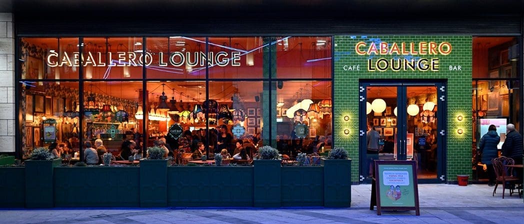 A vibrant street view of Caballero Lounge café and bar at dusk, with large windows revealing people dining inside. The entrance is framed by green tiles, illuminated signage, and features the name Caballero above the doors and windows.