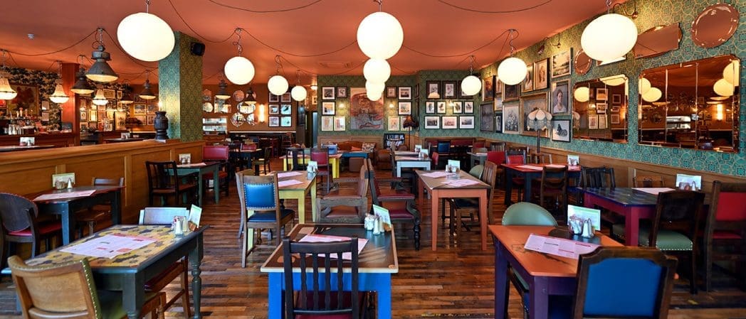 A cozy, eclectic restaurant interior with colorful mismatched chairs, wooden tables set with menus, round globe lights, and walls adorned with framed pictures and mirrors creates a warm Caballero-inspired charm.