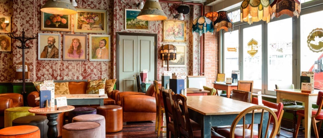 A cozy café with mismatched vintage furniture, colorful lampshades, and framed paintings of caballos on patterned wallpaper. Sunlight streams in through large windows, creating a warm, inviting atmosphere.