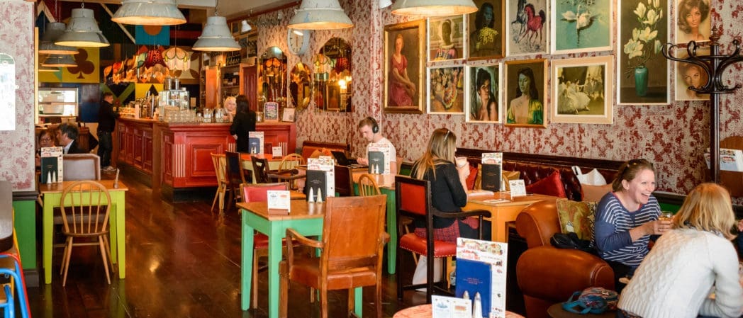 A cozy, eclectic café with mismatched chairs and colorful tables. People are seated and chatting. The walls are decorated with portraits, floral paintings, and a striking caballo mural. Warm lighting hangs from the ceiling, creating a welcoming atmosphere.