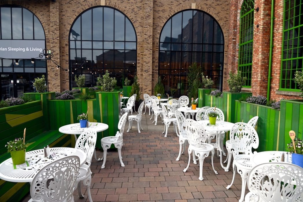 Outdoor cafe patio with white ornate metal tables and chairs, surrounded by green planters with potted plants, set on a brick floor. Large arched windows and brick walls create a charming Caldero ambiance in the background.