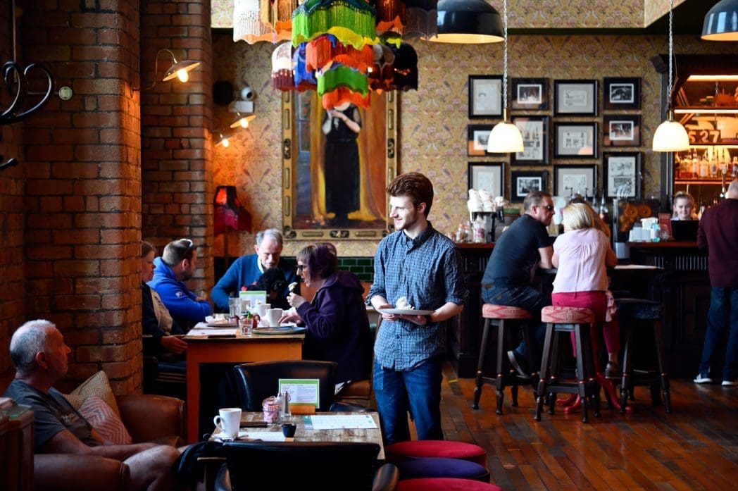 A server carries plates through a cozy, warmly lit café with patrons sitting at tables and the bar. Colorful hanging lamps, framed art, and a decorative caldero create a lively and inviting atmosphere.