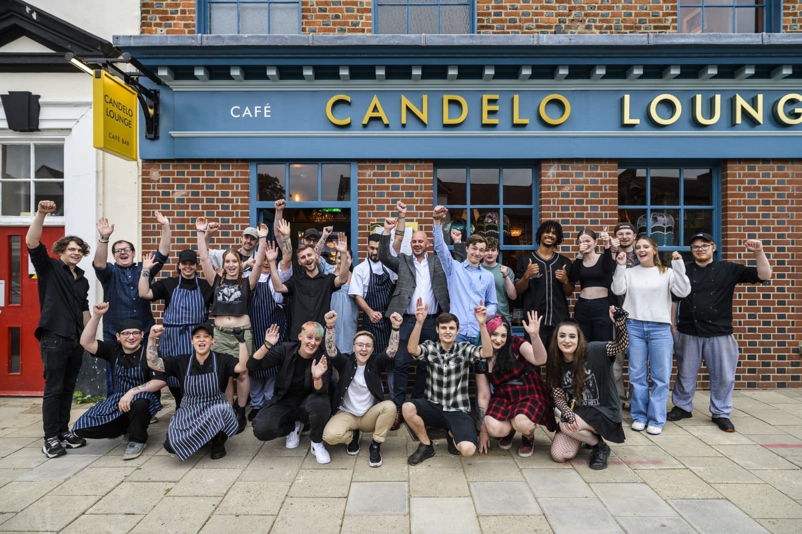 A large group of people pose and cheer with raised arms outside Candelo, standing on a sidewalk in front of the brick café building with large windows.