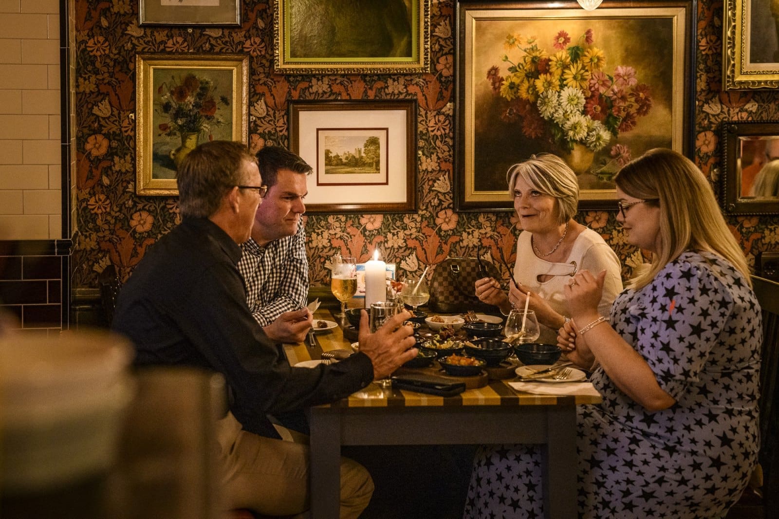 Four adults sit around a restaurant table at Candelo, enjoying food and conversation. The background features framed paintings and patterned wallpaper, creating a cozy, warmly lit atmosphere.