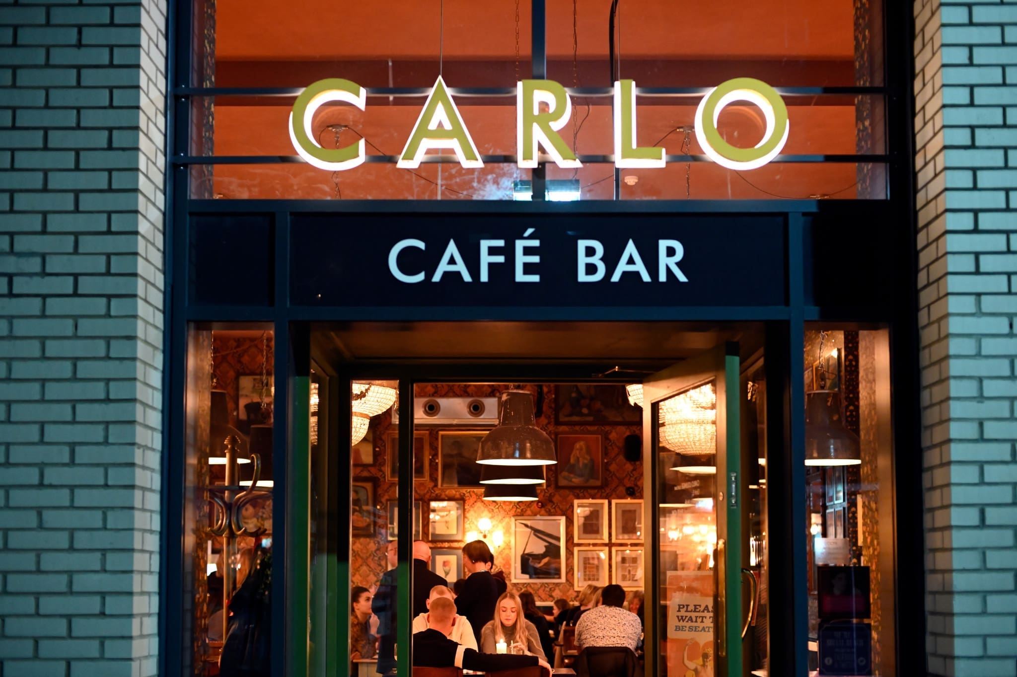 The photo shows the entrance of "Carlo Café Bar" at night, with Carlo’s warm lighting inside and several people sitting at tables, enjoying food and conversation. The interior walls are decorated with framed pictures.