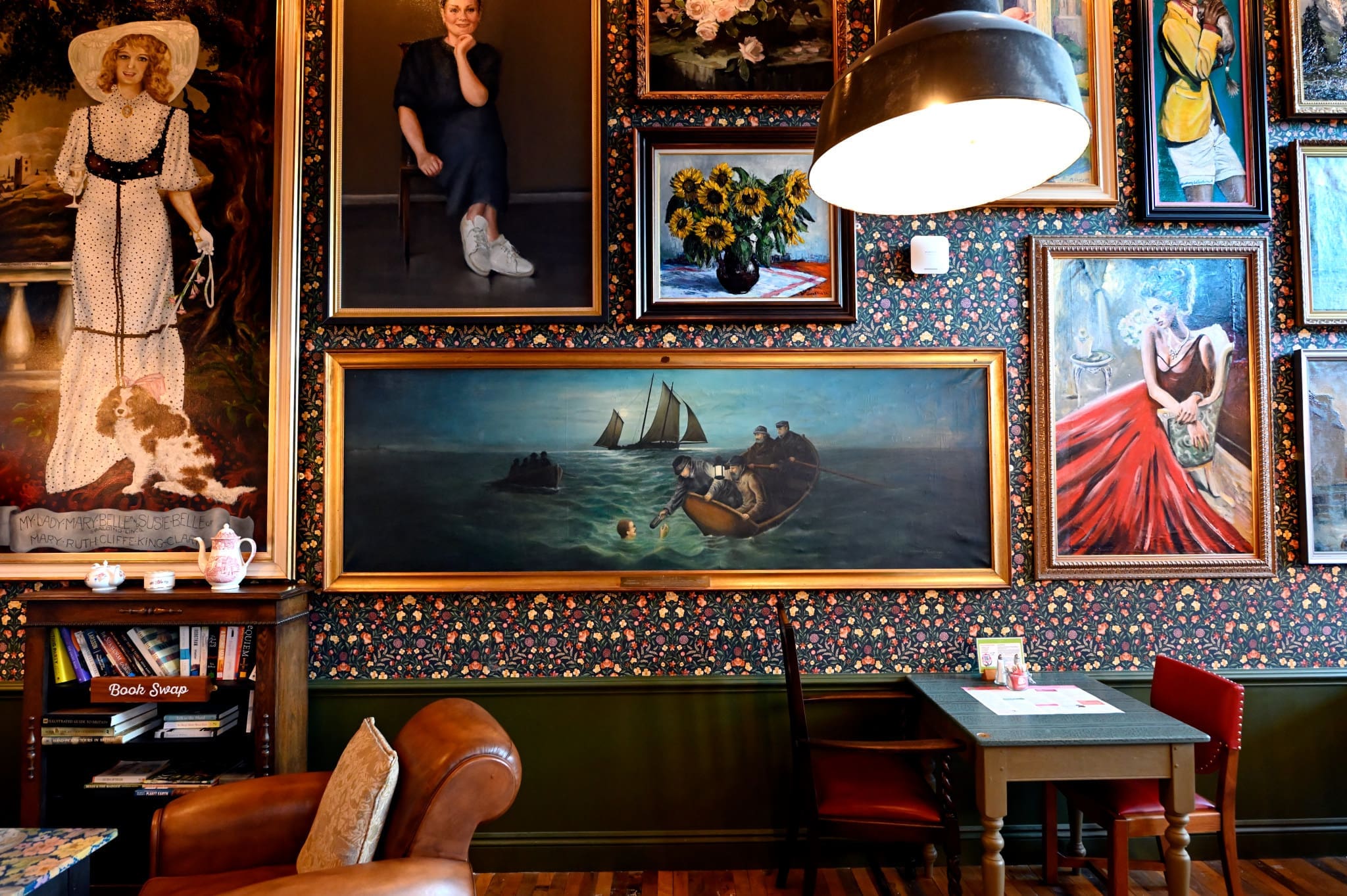 A cozy cafe with floral wallpaper, a leather armchair, green table, and walls adorned with Carlo’s framed paintings—portraits, sunflowers, and a seascape with boats. A large lamp hangs above the table.