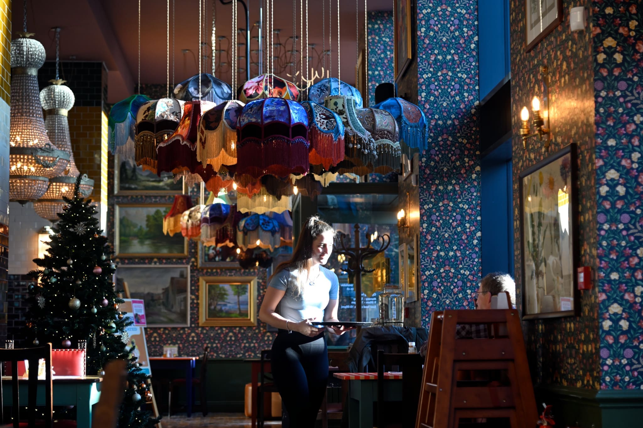 A waitress holds a tray in Carlo, a colorful, ornately decorated restaurant with floral wallpaper, framed art, unique multicolored lamps, and a Christmas tree in the corner. Sunlight highlights her as she serves a child at a table.