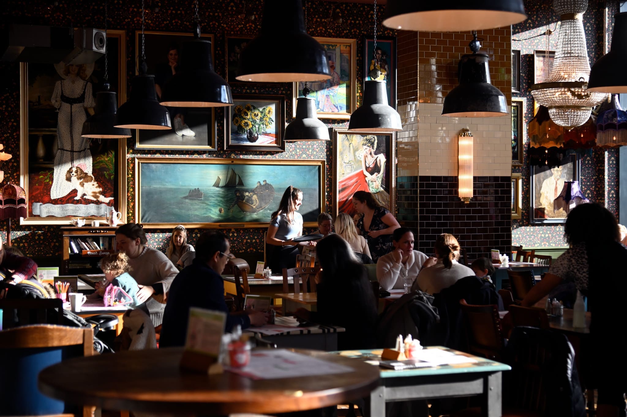 A lively restaurant with many people dining and chatting. Sunlight streams through the windows, illuminating eclectic wall art and paintings. Carlo, a server, stands near the center, taking an order from a table.