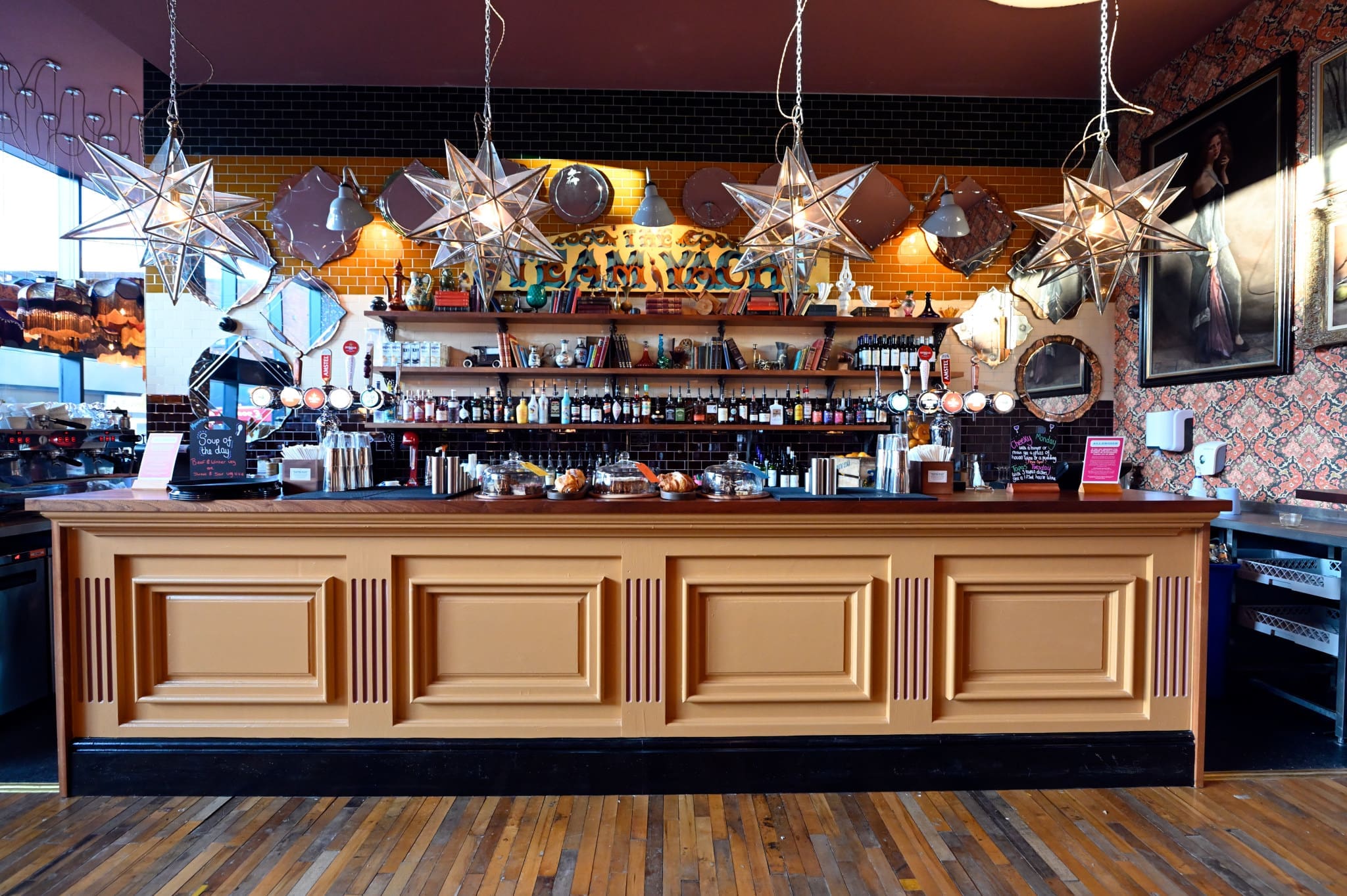 A stylish bar with a tan paneled counter, hanging star-shaped lights, various bottles and glasses on shelves, pastries on display, eclectic decor including framed art and wall patterns—perfect for a Carlo-inspired evening out.