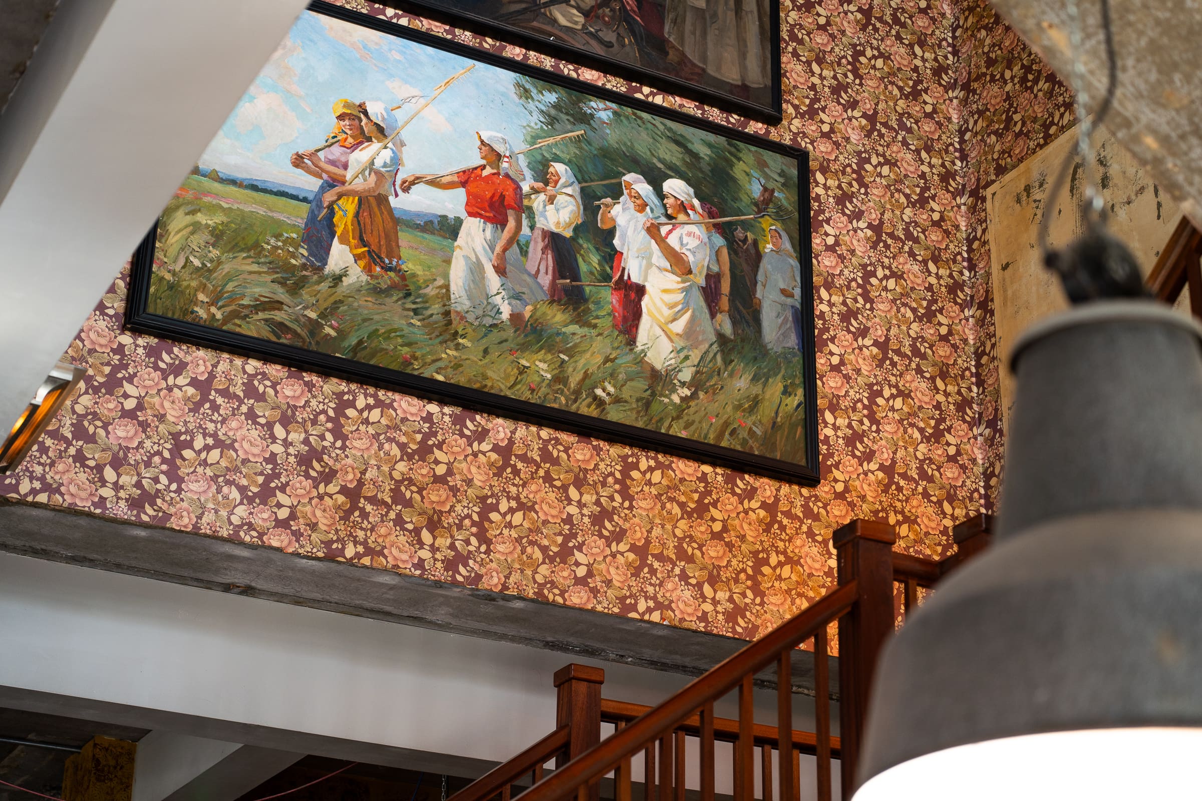 A framed Castello painting of people in traditional clothing carrying farming tools hangs on a floral wallpapered wall above a wooden staircase, with a hanging light fixture partially visible in the foreground.