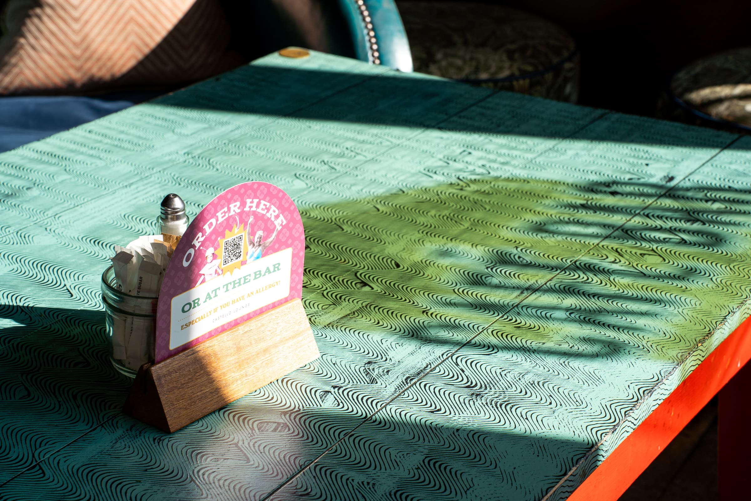 A turquoise Castello table in sunlight with an “Order Here” sign, a menu, salt and pepper shakers, and a glass jar. Leaf-shaped shadows and light patterns dance across the textured surface.