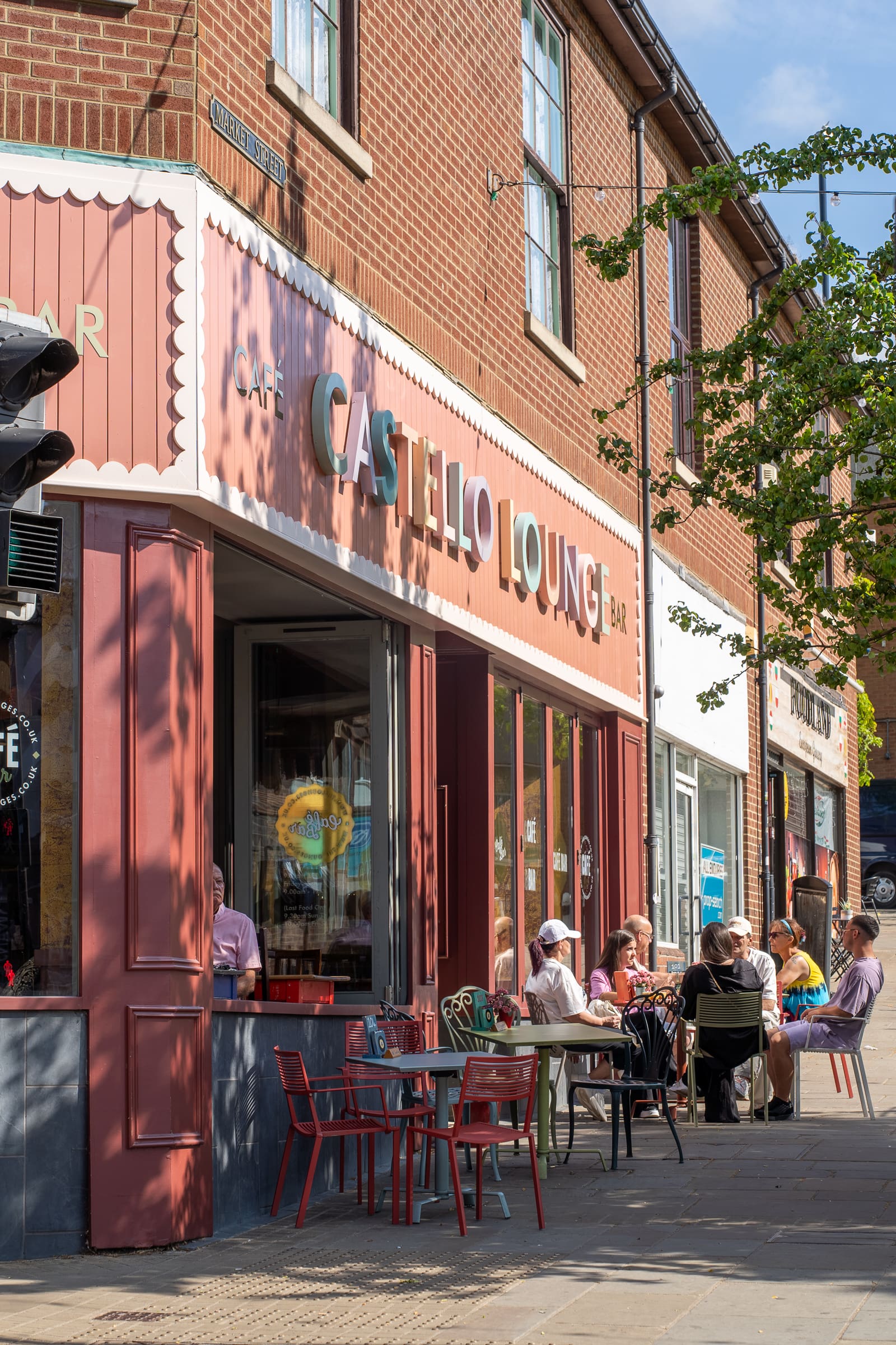 People sitting at outdoor tables in front of Castello Lounge café and bar on a sunny day, with a tree providing some shade and neighboring shops visible along Castello Street.