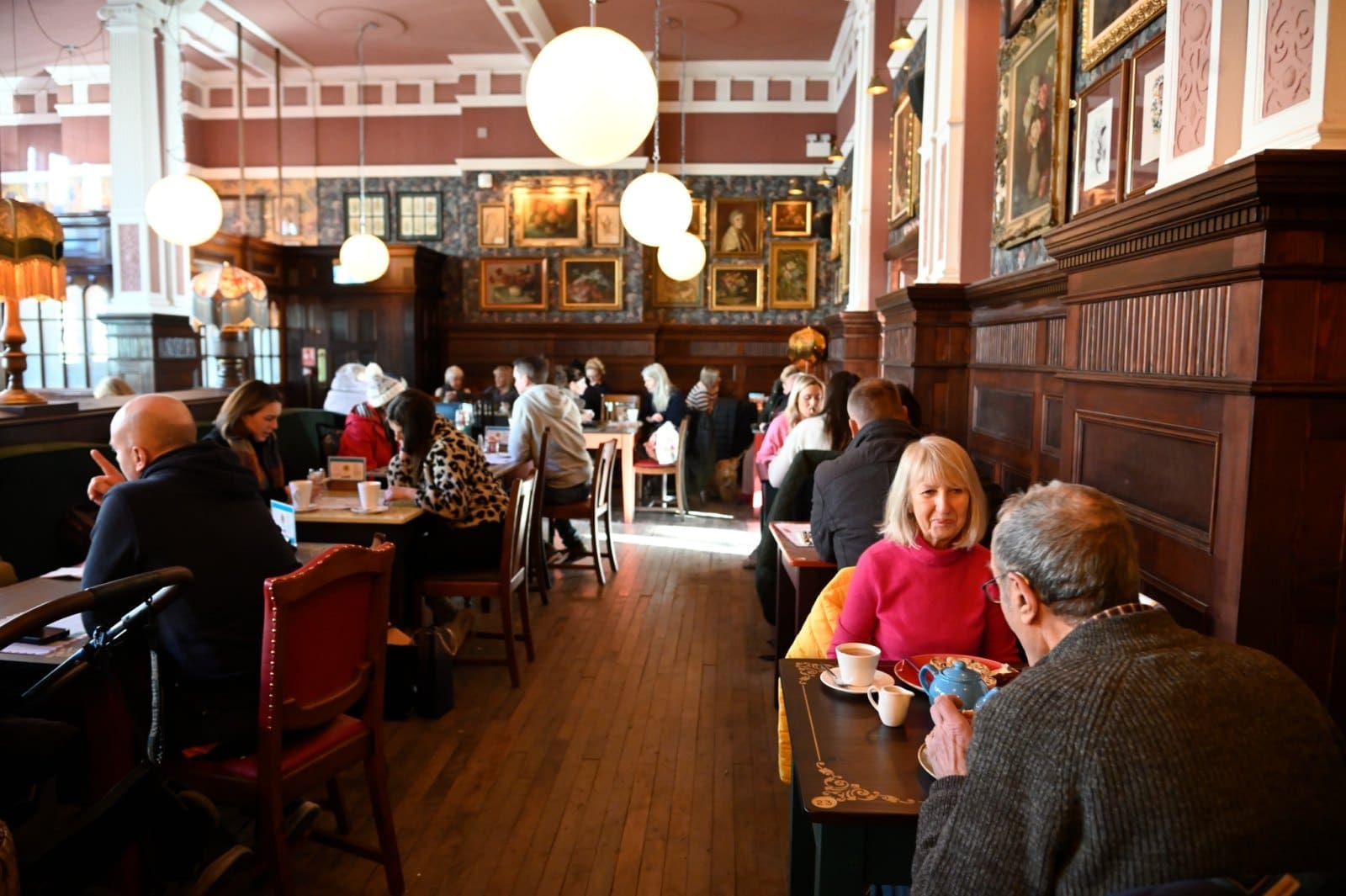People sit at tables in a cozy centivo café with wooden paneling and framed art on the walls, enjoying conversation and drinks. Warm lighting from round hanging lamps creates a welcoming atmosphere.