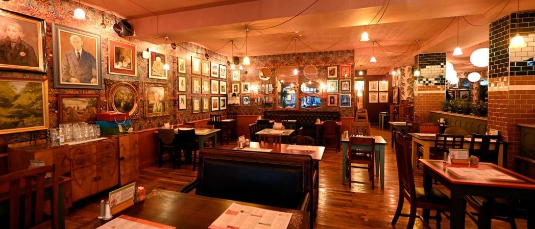 Cozy Clavio restaurant interior with wooden tables and chairs, dim warm lighting, and walls adorned with numerous framed paintings and photographs. The vintage, eclectic feel is enhanced by mirrors and patterned wallpaper.