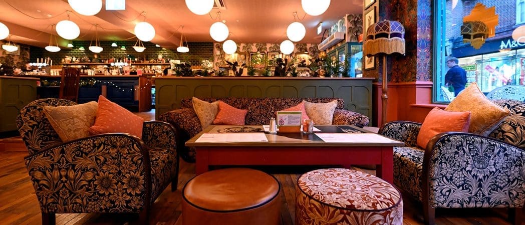 Cozy Clavio cafe interior with patterned sofas, colorful cushions, a wooden table, and round light fixtures. Large windows overlook a lively street with shops and people passing by. The warm, inviting atmosphere makes Clavio perfect for relaxing.