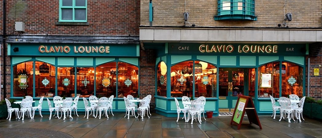 Clavio Lounge is a teal-fronted café and bar with large windows, warm interior lighting, and white ornate outdoor tables and chairs arranged on the wet pavement.