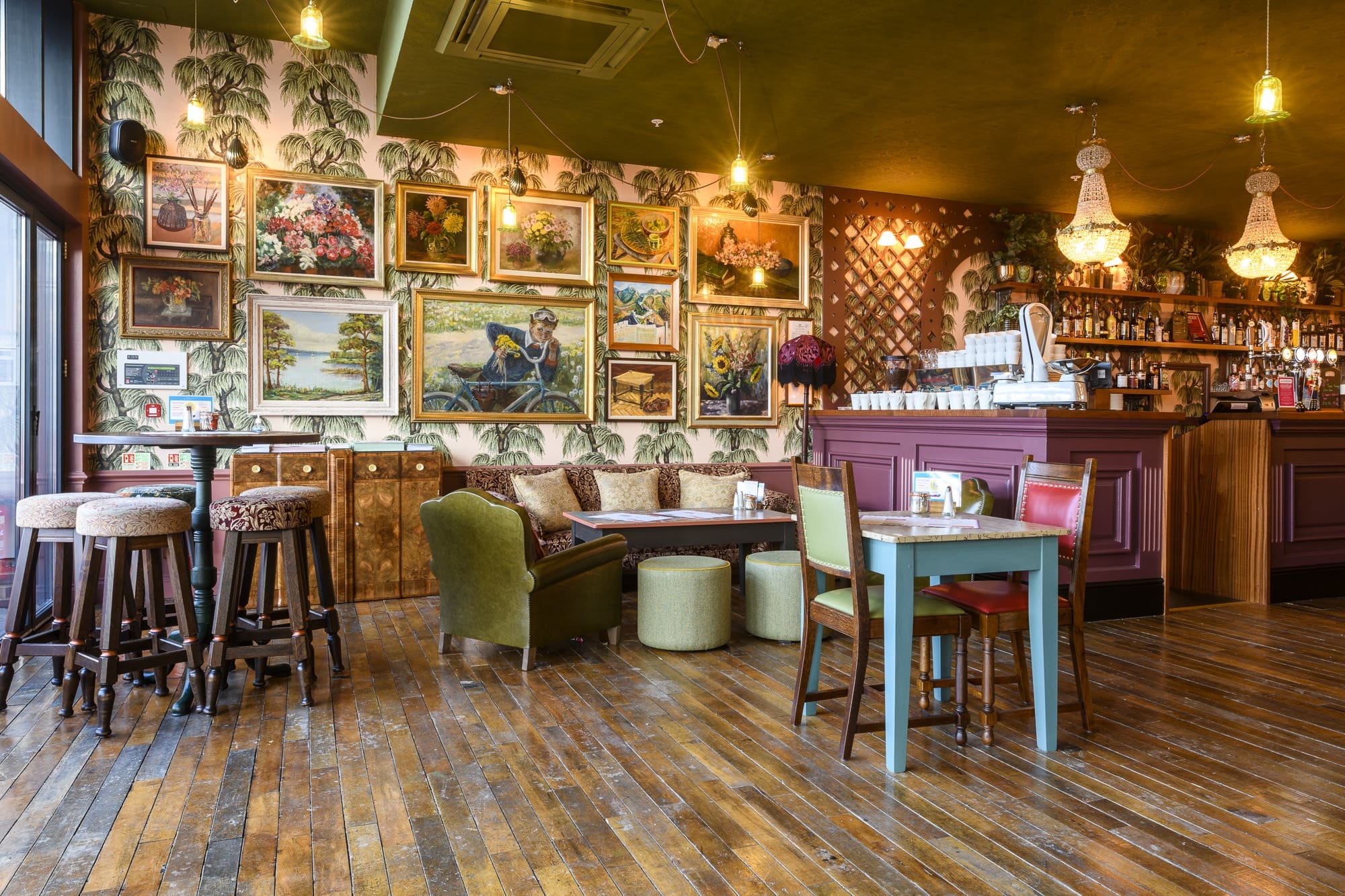A cozy café with wooden floors, vintage-style furniture, and a wall of framed paintings. The bar area is to the right, coltello neatly arranged beside the coffee machine on the counter beneath sparkling chandeliers.