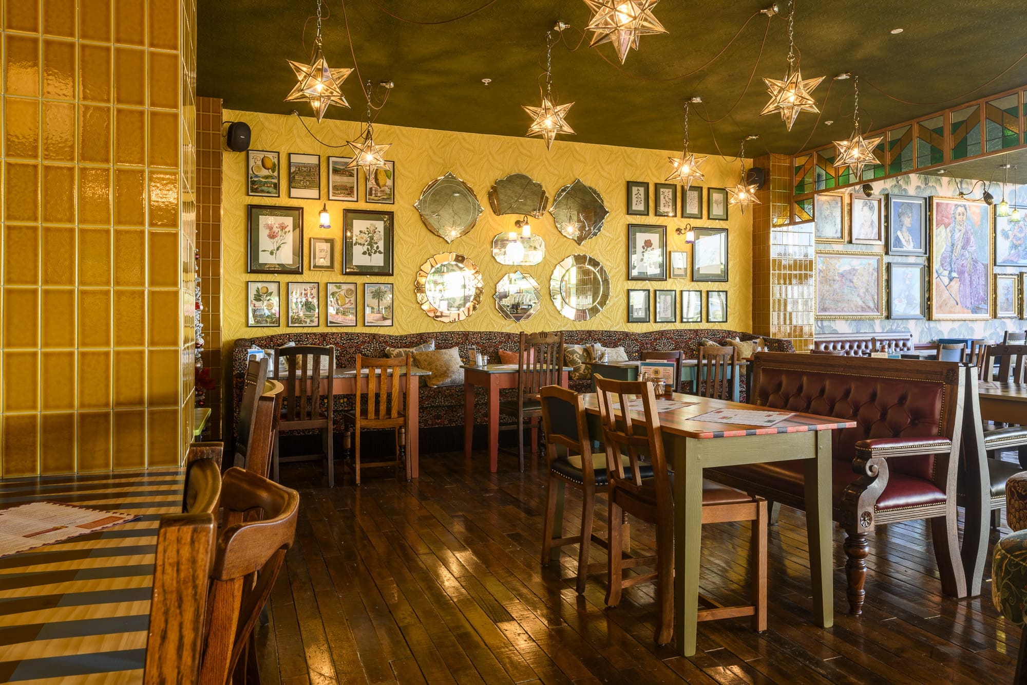 A cozy, eclectic restaurant interior with wooden floors, vintage chairs, star-shaped pendant lights, and walls decorated with assorted mirrors and artwork. Tables are set with gleaming coltello and the inviting atmosphere welcomes you in.